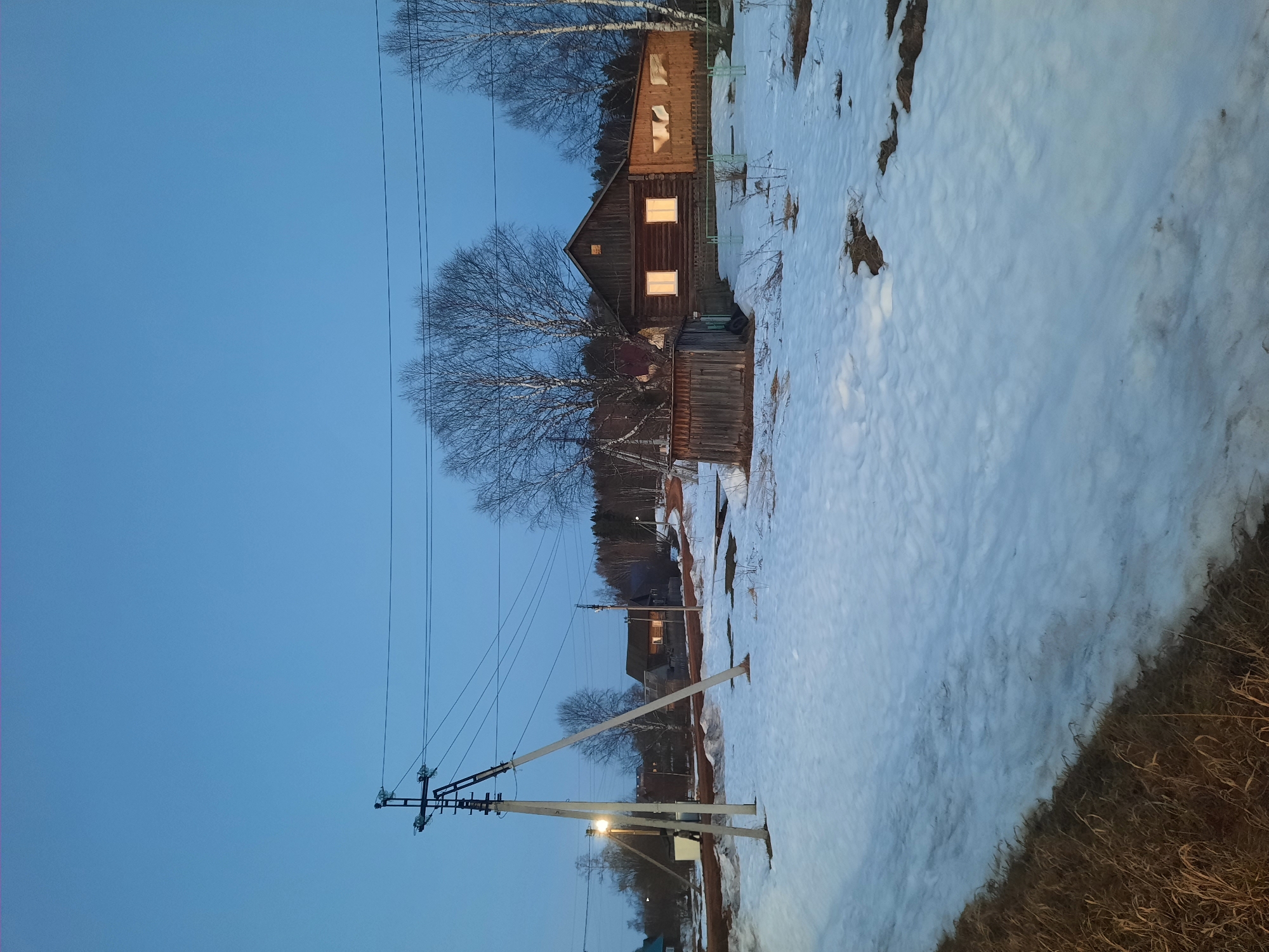 The image shows a rural scene in Krasnogorskoye, Russia, at dusk. Patches of snow cover the ground. A wooden house with illuminated windows stands on the right side of the frame. Bare trees surround the house. Power lines and poles extend from the left side of the image, crossing the sky. The sky is a clear, deep blue. Grass is visible in the foreground, peeking out from under the snow.