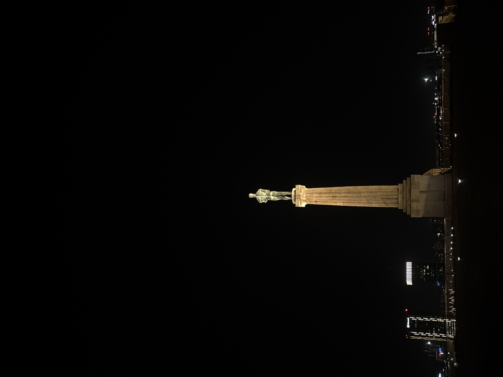 At night, a statue atop a tall pillar is situated in a city, with Belgrade, Serbia, being visible along the horizon. The statue is of a human figure, possibly a man, standing tall on the pillar. The pillar itself is illuminated. The cityscape includes buildings and lights along the waterfront. The background is dark, highlighting the statue and the lighted structures in the urban area.
