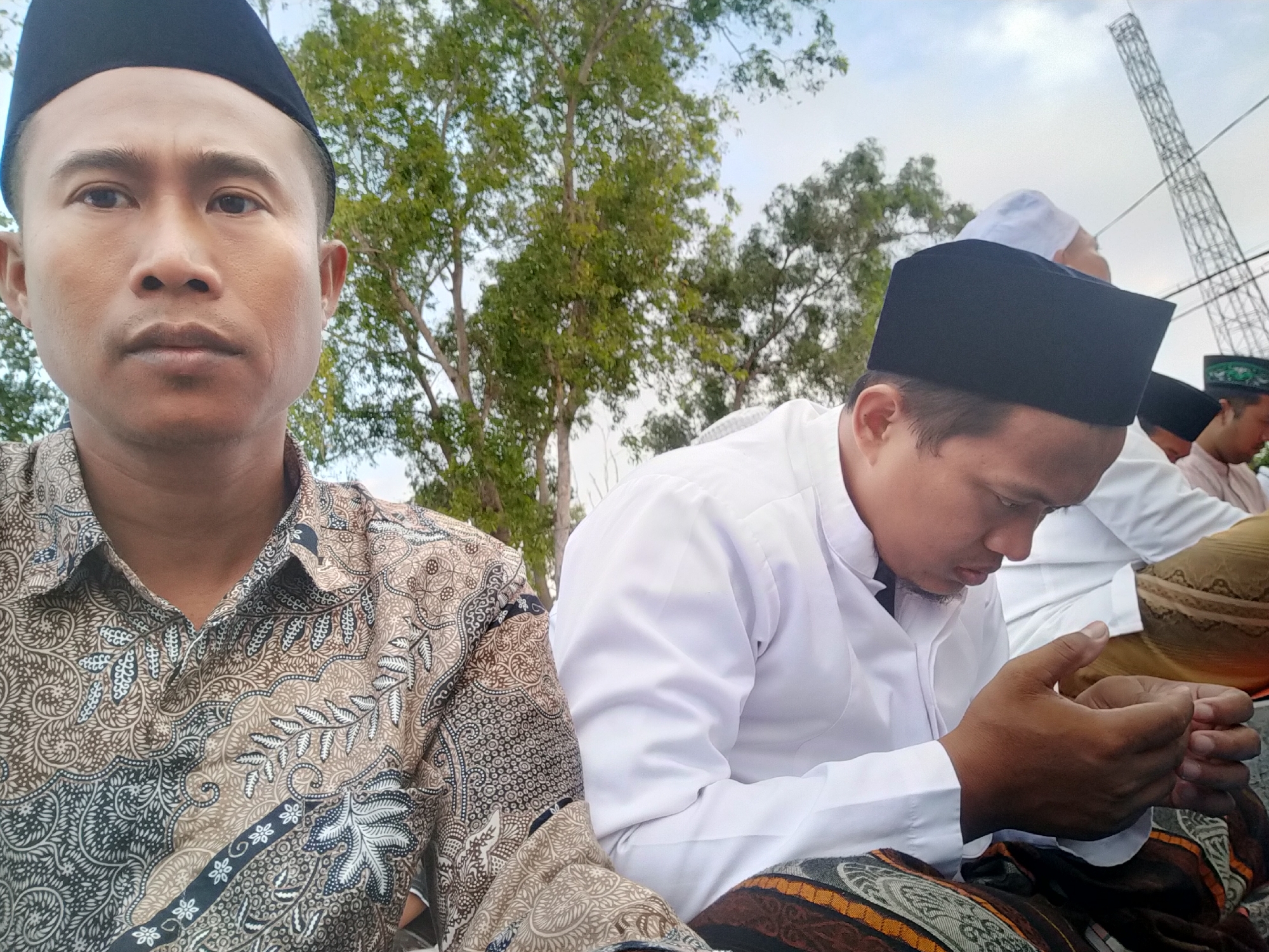 The image, taken outdoors, shows four Muslim men, three of whom are visible from the chest up. The man in the foreground on the left is looking directly at the camera, his face is center-frame. He is wearing a black peci (a brimless, rounded cap) and a patterned brown and white shirt. To his right, another man wearing a black peci and a white shirt is looking down and holding something in his hands, which are clasped. Behind him, another man wearing a white peci and a white garment is partially visible, looking to the right. A fourth man, in the background to the right, is also wearing a peci and a white shirt, and is looking downwards. The background consists of trees and a large, metal tower structure against a cloudy sky. The scene appears to be set in Kranji, Indonesia, given the context of the image.