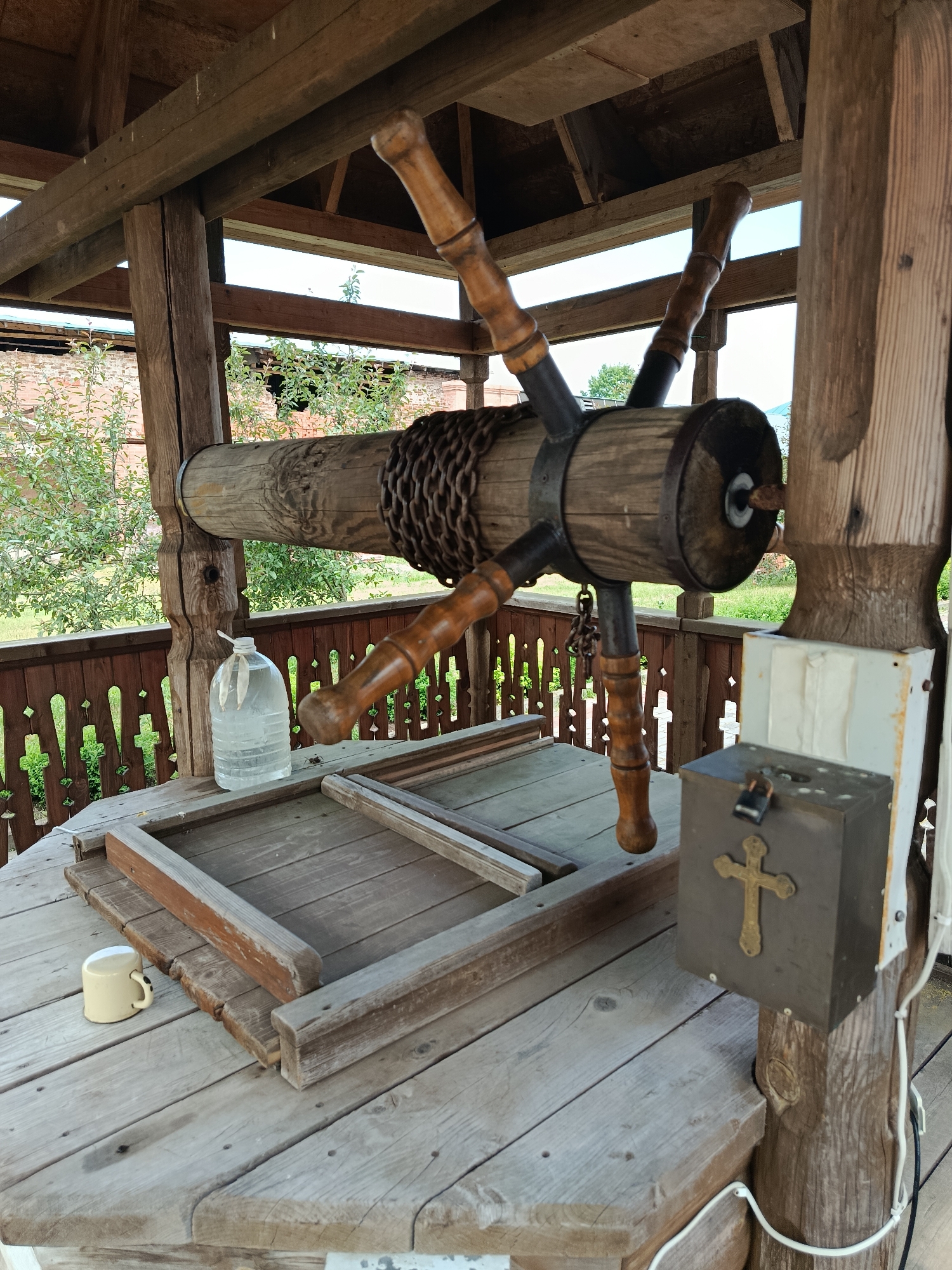 This is a photograph of an outdoor well in the village of Radovitsy, Russia. The well has a wooden structure with a rope and pulley system for drawing water. A large plastic bottle filled with water is visible on the well's wooden platform, along with a small metal mug and a wooden frame. A dark metal box with a padlock and a golden cross is mounted on one of the well's support posts. The background shows trees and a brick building. The overall scene suggests a rustic and traditional setting.