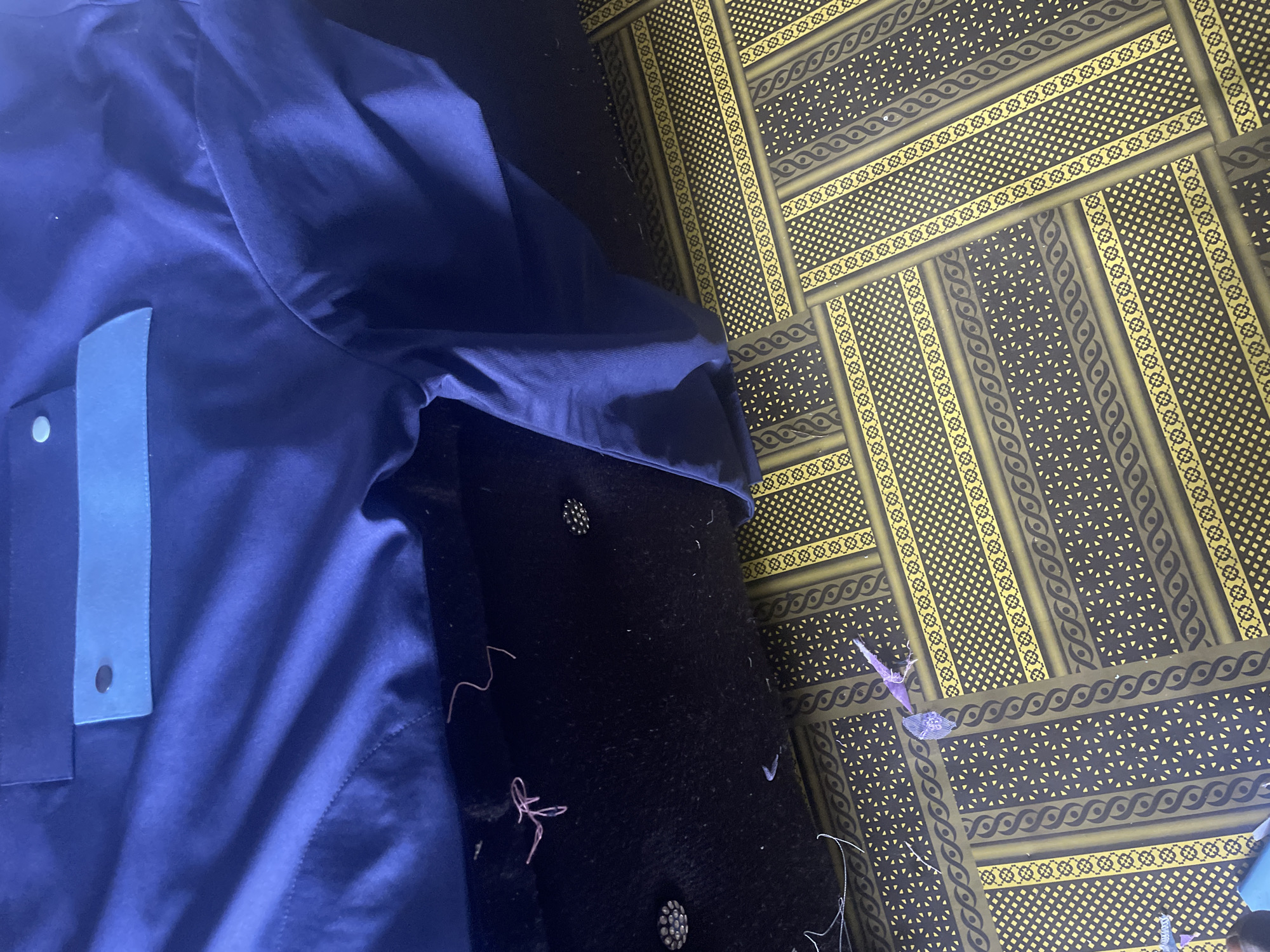The image shows a close-up view of a dark blue garment draped over a black textured surface. The garment features a light blue pocket with a button closure. The black surface below appears to be some form of fabric or upholstery, with loose pink threads scattered on it. The lower portion of the image displays a patterned surface with yellow and black geometric designs, resembling a floor covering or textile. There are no people or explicit actions visible. The scene appears to be an indoor setting, possibly a workshop or a domestic space. It is not possible to determine the location within Jauro Yinu, Nigeria based solely on the visual information provided.