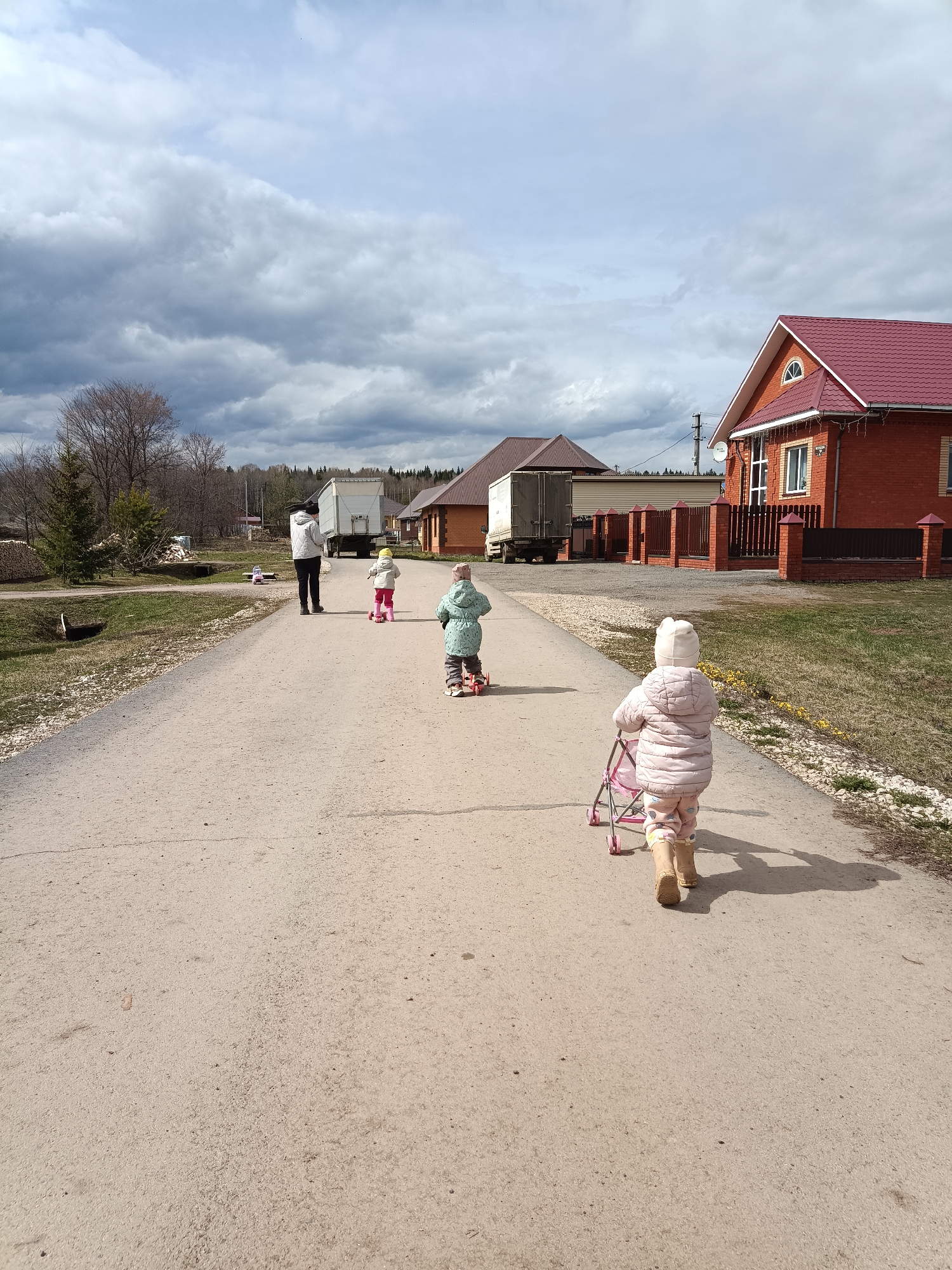 A medium, eye-level shot depicts a group of four individuals on a lightly textured, concrete road in what appears to be a rural residential area. The sky is overcast with grey storm clouds. From front to back, the individuals consist of: a young child walking away from the camera and pushing a pink toy stroller; a young child on a scooter; a young child on a scooter wearing a yellow hat; and an adult standing still. The young children all wear winter coats. Two large trucks are parked parallel to the road. Adjacent to the road are small patches of grass, some with sparse yellow flowers. A red brick house with a red roof is situated on the right side of the road. The location is Barda, Russia.