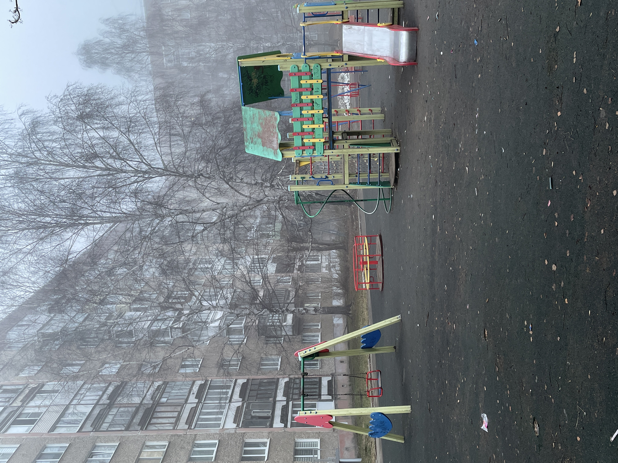 The image depicts a playground in Izhevsk, Russia, on a foggy day. The playground features a slide, a climbing structure, a swing set, and a rotating seat. The ground is asphalt, covered in scattered leaves and debris. A large, multi-story apartment building rises in the background, partially obscured by the fog. A bare tree stands beside the playground. No people are visible.