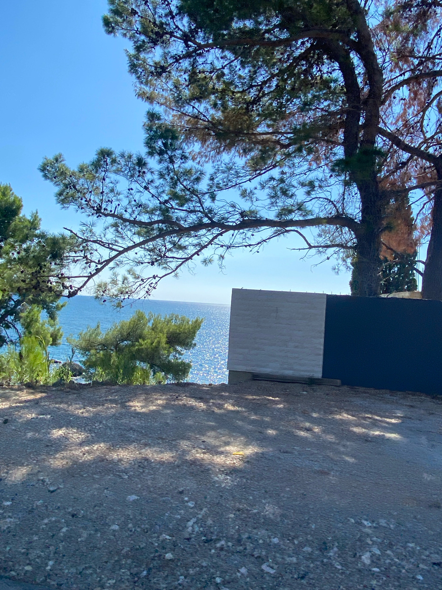 The image depicts a scenic view of the Adriatic Sea, as seen from an elevated vantage point, likely within the city of Bar, Montenegro. In the foreground, a coarse gravel path is visible, with dappled sunlight casting shadows from the overhead pine trees. These pine trees, with their needles and cones, frame the upper portion of the image. Beyond the path, a section of a modern fence or wall structure is present. This structure is divided into two distinct parts: a left section with a light grey, textured surface resembling brickwork, and a right section covered in dark material, possibly black.
The middle ground showcases the glittering blue sea, with sunlight reflecting off its surface, creating a shimmering effect. Lush green vegetation, including smaller pine trees and reeds, grows along the coastline, adding to the natural beauty of the scene. The horizon line, where the sea meets the clear blue sky, is visible in the distance. The overall atmosphere is one of a bright, sunny day, offering a serene coastal landscape. No people or specific structures of Bar are prominently identifiable, but the presence of pine trees and the sea strongly suggests a coastal location consistent with Bar.