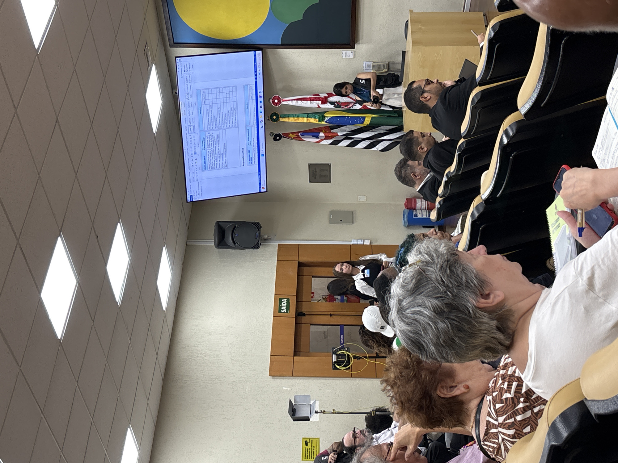 The image captures an indoor scene of a meeting in what appears to be a public or governmental setting. A group of people, mostly seated, are looking towards a large screen displaying a document. Flags of Brazil, São Paulo and other political entities are behind the stage. The meeting is potentially related to the city of São Paulo, Brazil, due to the flag of São Paulo present. A speaker is standing near the exit to the stage, presumably addressing the crowd. A camera man is near the flags taking photos. The room is lit by diamond shaped ceiling lights.