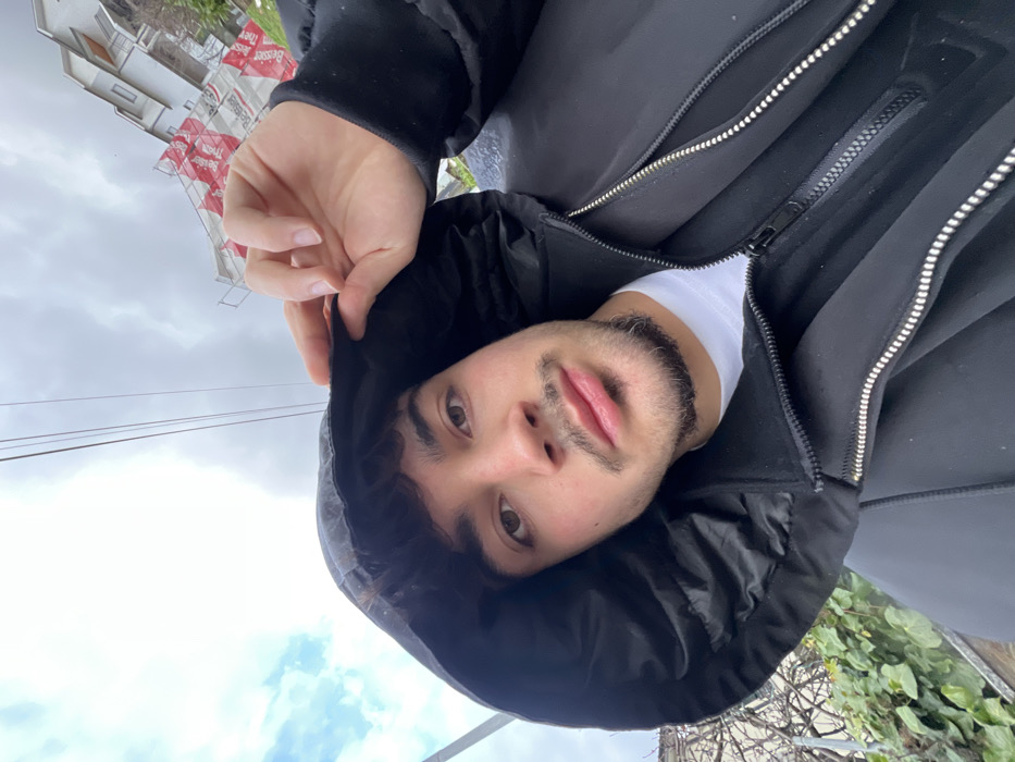 A young man is prominently featured in a close-up, outdoor portrait, likely a selfie. He is looking directly at the camera with a neutral and calm expression. His head and upper torso fill most of the frame.
He has dark, curly hair, brown eyes, and full lips. A short beard, appearing dark with some lighter or grey hairs mixed in (particularly visible around his chin and mustache), frames his jawline. He is wearing a dark, possibly black, hooded puffer jacket. A white t-shirt is visible underneath the jacket, peeking out at the collar and chest. His right hand is visible, holding onto the edge of his hood, slightly pulling it up around his head. The jacket features visible zippers, suggesting a layered design.
The background, under an overcast sky, suggests an urban or suburban setting. Light grey clouds fill much of the sky. In the left mid-ground, parts of white and grey buildings are visible. In front of or attached to these buildings are several red and white banners. Partially visible text on these banners includes what appears to be German words: "Besser" (meaning "better"), "Rieger" (a surname), and "ihrer" (meaning "their" or formal "your"), suggesting an advertisement, possibly for real estate or a local business. Thin, dark lines, likely power or telephone lines, stretch across the sky in the middle distance. In the lower right corner of the image, there is a patch of green foliage, possibly ivy or a bush, indicating the presence of plants.
The lighting is soft and diffuse, consistent with an overcast day, and there are no harsh shadows. The overall scene captures a candid moment in an everyday outdoor environment.