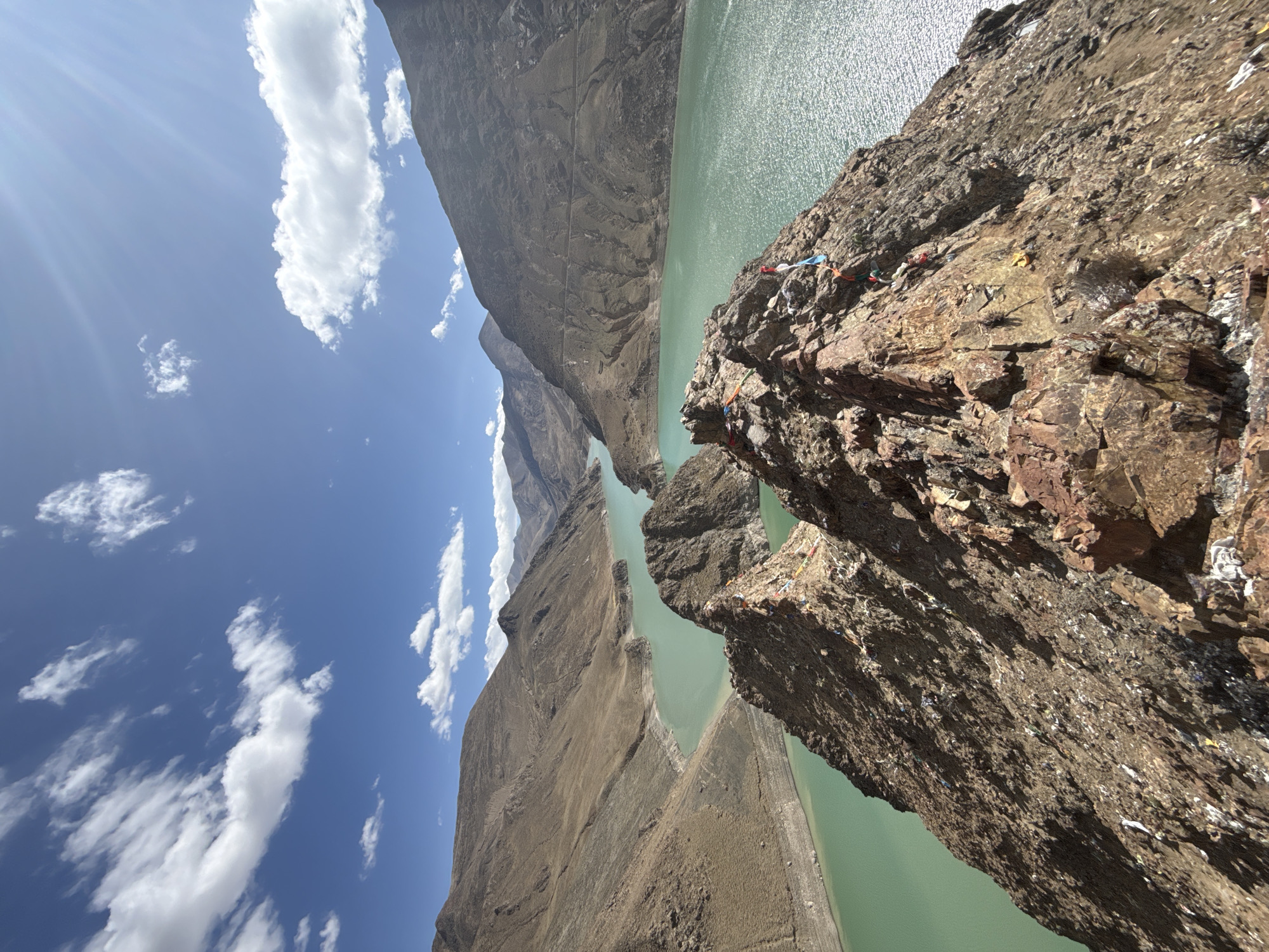 The image shows a view overlooking a canyon or gorge in Gyantse County, China. A large body of green-tinted water fills the canyon floor. On either side, steep, rocky cliffs rise, the one in the foreground displaying a rough, layered texture. Prayer flags are attached to the rocks near the edge. The sky above is blue with scattered white clouds and visible sun rays. The image is taken from a high vantage point, likely from one of the cliffs.