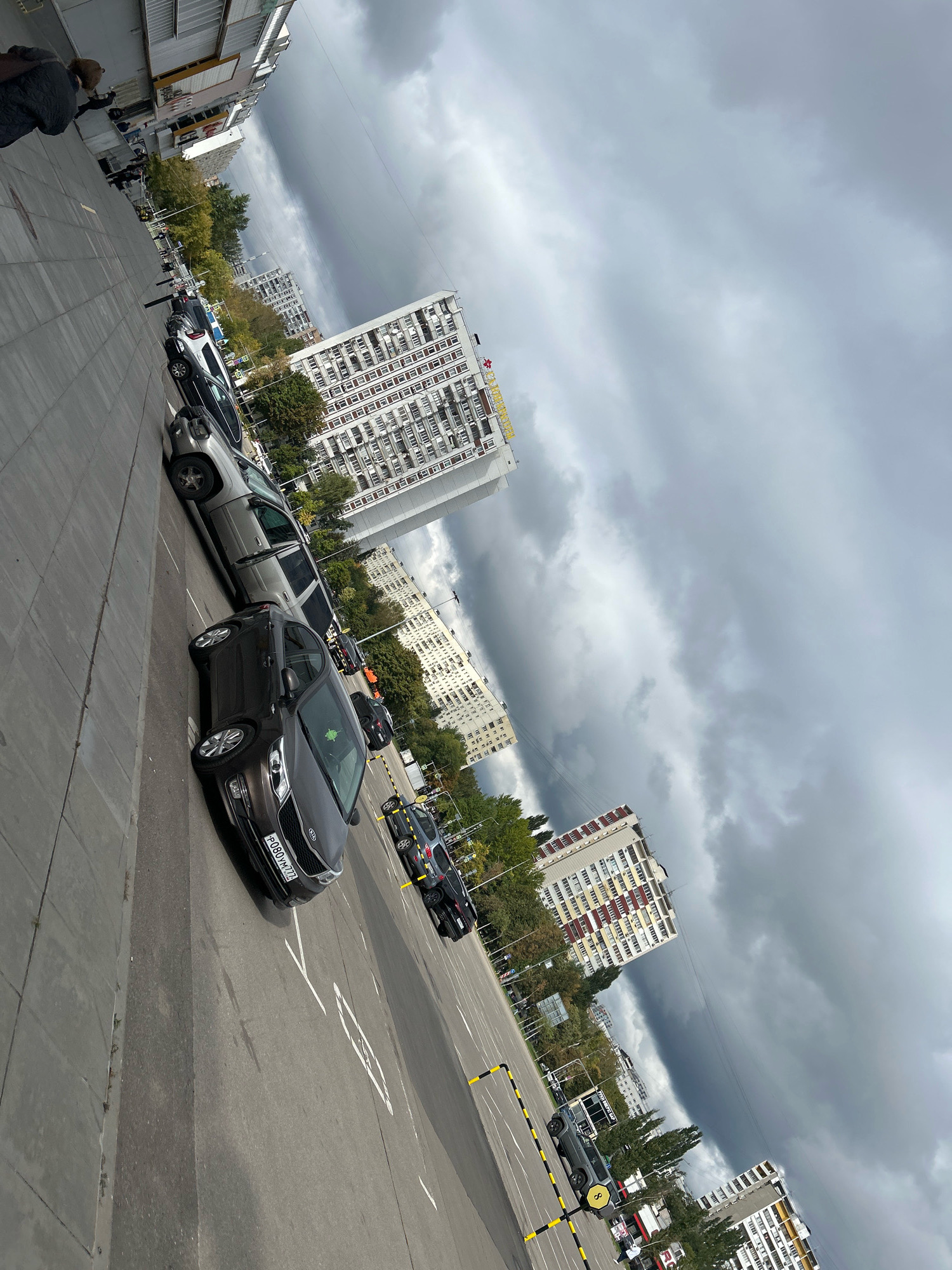 The image depicts a street scene in Moscow, Russia, under a cloudy sky. A row of cars is parked along a sidewalk adjacent to a street. In the background, tall apartment buildings line the street, interspersed with trees. The perspective is from street level, looking forward and slightly upwards. The image is rotated, with the bottom of the frame appearing on the left.