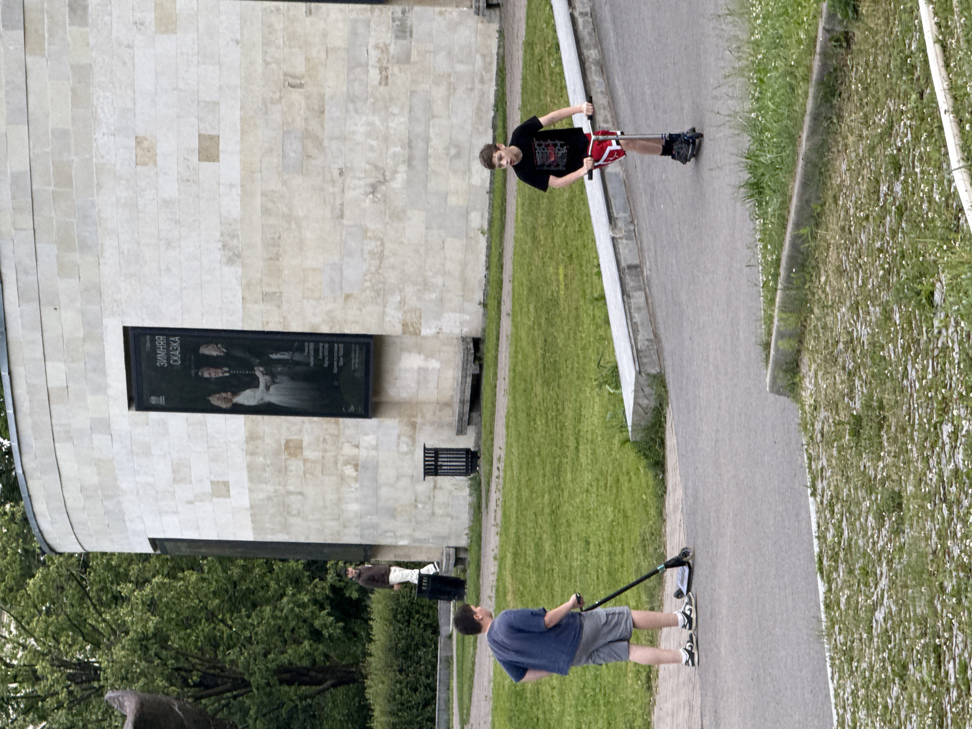 Two young men are seen in the media file, on a sunny day in what appears to be an urban park or plaza in Saint Petersburg, Russia. The scene is dominated by a modern building with a light-colored, tiled facade. The building has a large, dark poster displayed on its exterior.
One young man is riding a scooter, wearing a black t-shirt with red accents and red shorts. He is looking forward, and the scooter appears to be moving. The second young man is walking, holding onto the handlebars of a scooter that is parked on the ground. He is wearing a blue t-shirt and grey shorts.
The ground consists of a paved walkway adjacent to a well-maintained grassy area with scattered small white flowers. In the background, lush green trees suggest a pleasant, possibly spring or summer, day. The lighting indicates it is daytime. The overall mood seems casual and relaxed, with the individuals engaged in leisurely outdoor activity. The poster on the building has some visible text in Cyrillic script, reading "ЗИМНЯЯ СКАЗКА" (Zimnyaya Skazka), which translates to "Winter Fairy Tale."