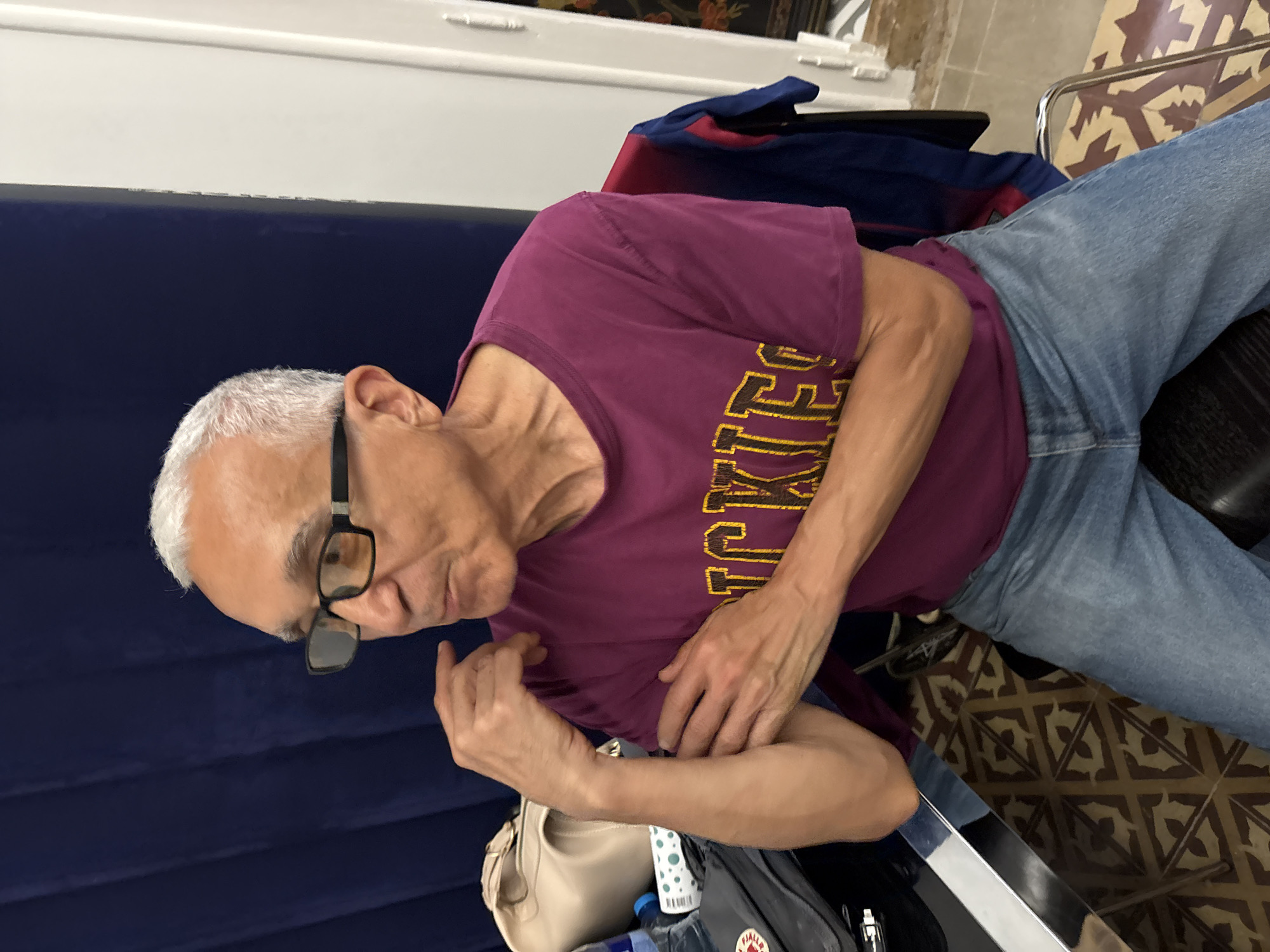 The image shows an older man with short gray hair, wearing glasses, a maroon T-shirt with yellow lettering, and blue jeans. He is seated in a chair. He is looking down, his hands resting on his lap. In the background, there's a blue curtain, a tiled floor with a geometric pattern, and a backpack is near his feet. While not conclusive based on the provided image alone, the tile work suggests this may be within a historic building in Barcelona, Spain.