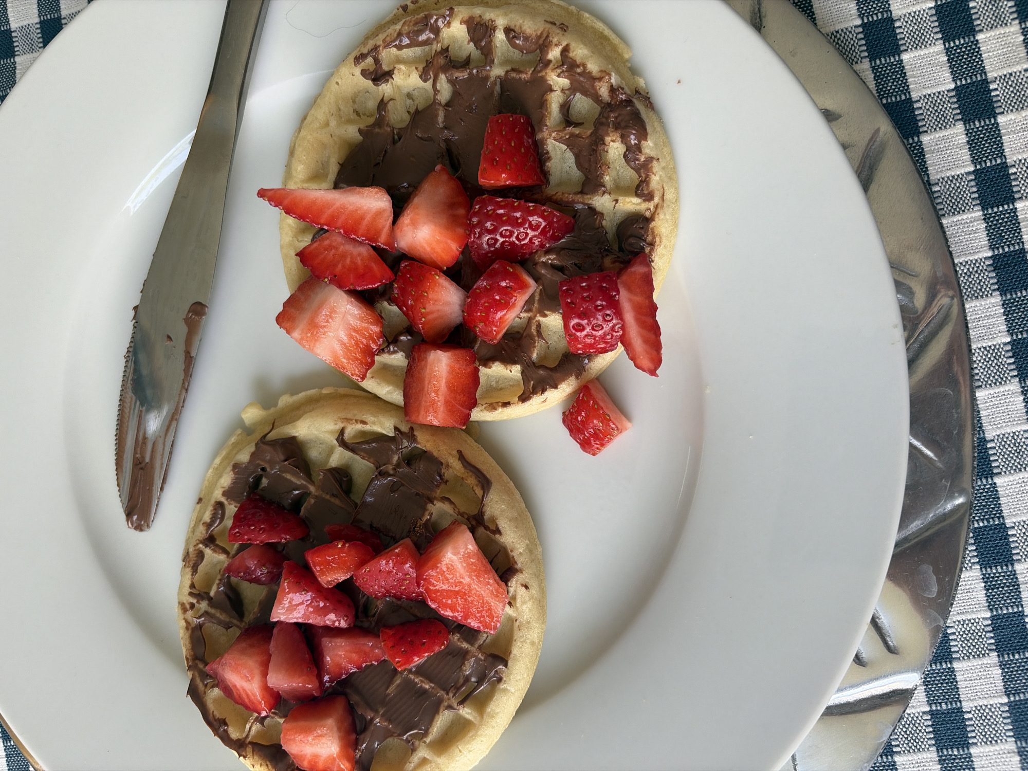 A close-up, top-down shot depicts a white plate holding two waffles topped with chocolate spread and diced strawberries, alongside a silver knife. The waffles are round and golden brown, with the chocolate spread smeared unevenly across their surfaces. The strawberries are bright red and cut into small pieces, scattered on top of the chocolate. The silver knife rests on the plate, its blade slightly coated with the chocolate spread. The plate is set against a blue and white checkered surface. The location is not specified.
