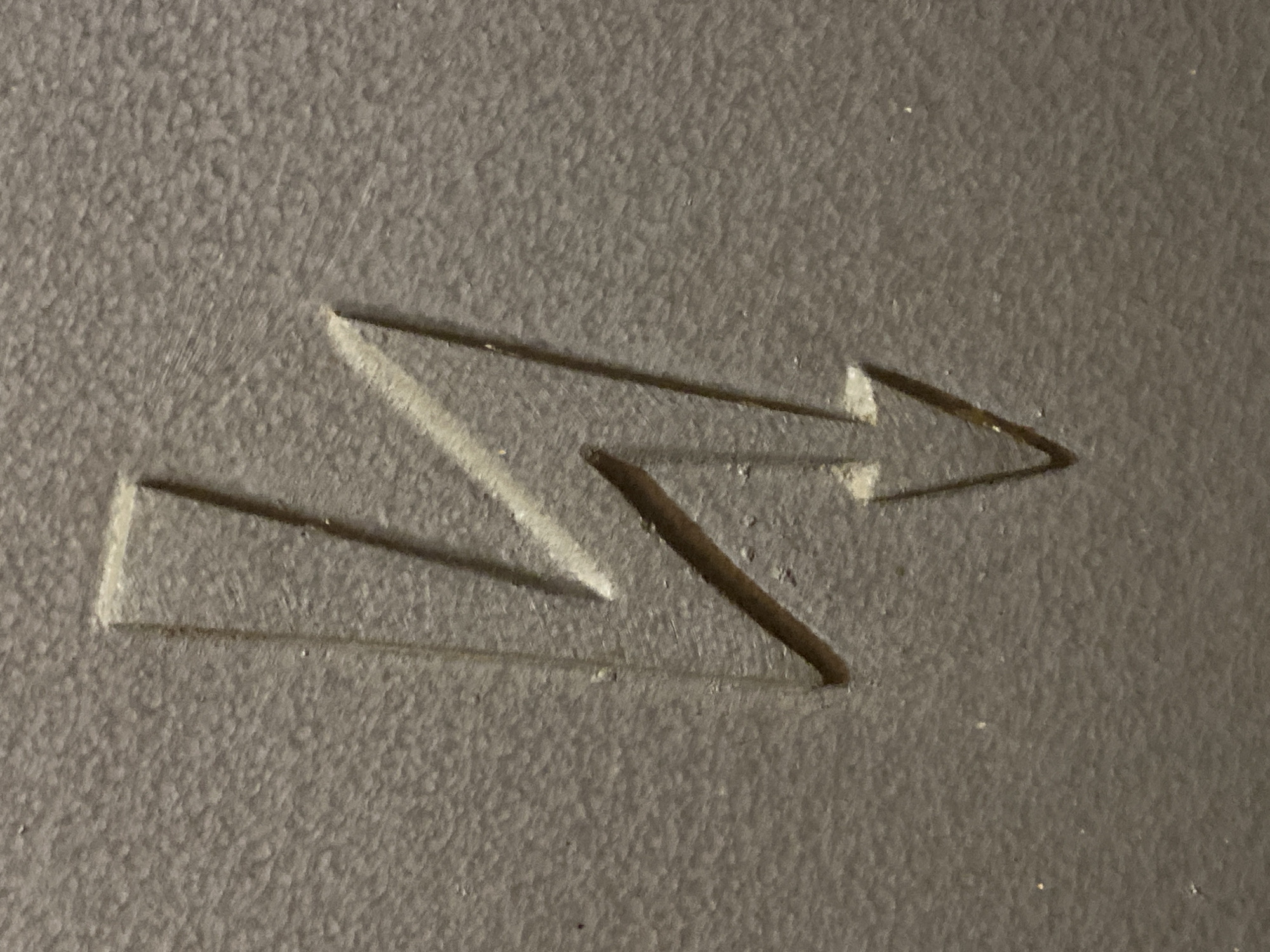 The image shows a stylized arrow symbol etched into a rough, grainy surface. The arrow is composed of angular lines, resembling a combination of the letter "S" and an arrow pointing to the right. There are no people, animals, or any other objects visible in the image besides the etched arrow and the material it's etched into. The environment is limited to the texture and color of the surface material. The image does not contain enough information to locate the specific place in Budva, Montenegro.