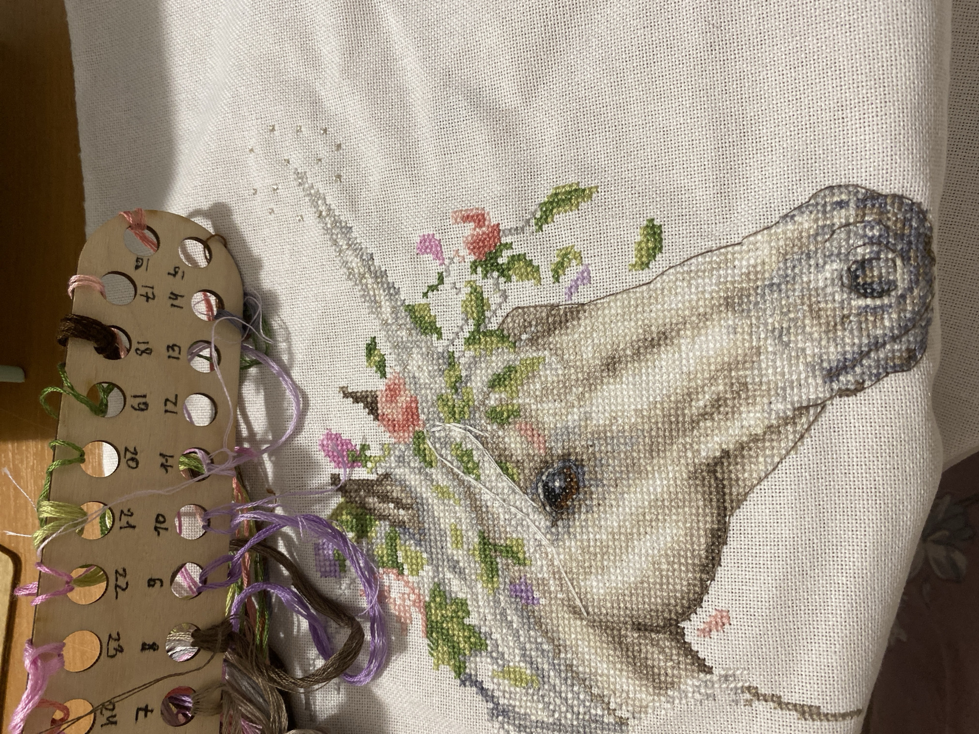 A close-up photograph shows a partially completed cross-stitch project of a unicorn's head, adorned with floral accents. The unicorn is primarily stitched in varying shades of gray and brown, giving it a realistic appearance. Flowers, in shades of pink and green, are woven into the mane and around the unicorn's horn. The stitches appear neat and uniform. A needle with thread is currently in use.
To the left of the cross-stitch is a wooden thread organizer. The organizer features numbered holes, each containing different colors of embroidery floss. The numbers are printed next to each hole, ranging from 16 to 24, but possibly higher. The floss colors include greens, purples, browns, and pinks.
The background of the image consists of the white fabric of the project, with some areas still unstitched. The lighting is diffused, creating soft shadows and highlighting the textures of the cross-stitch.