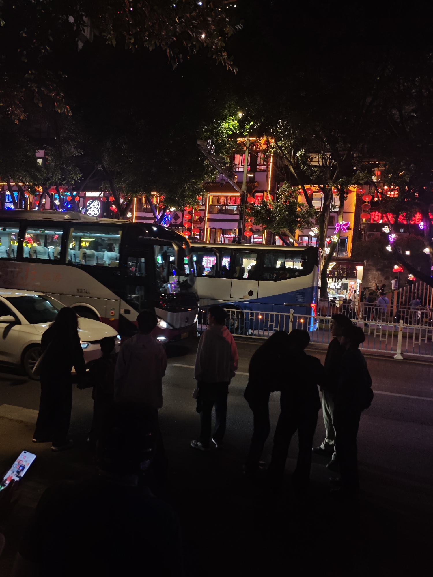 The image shows a street scene at night in what appears to be the Yuzhong District of Chongqing, China. The background reveals buildings adorned with red lanterns and decorative lighting. In the foreground, several people are grouped together on the sidewalk, some appearing to be looking toward the buildings across the street. A white car and two buses are parked along the road. One person is holding a smartphone, visible in the lower left corner. A white metal fence separates the sidewalk from the street in some sections. The overall environment suggests a lively atmosphere within an urban setting.