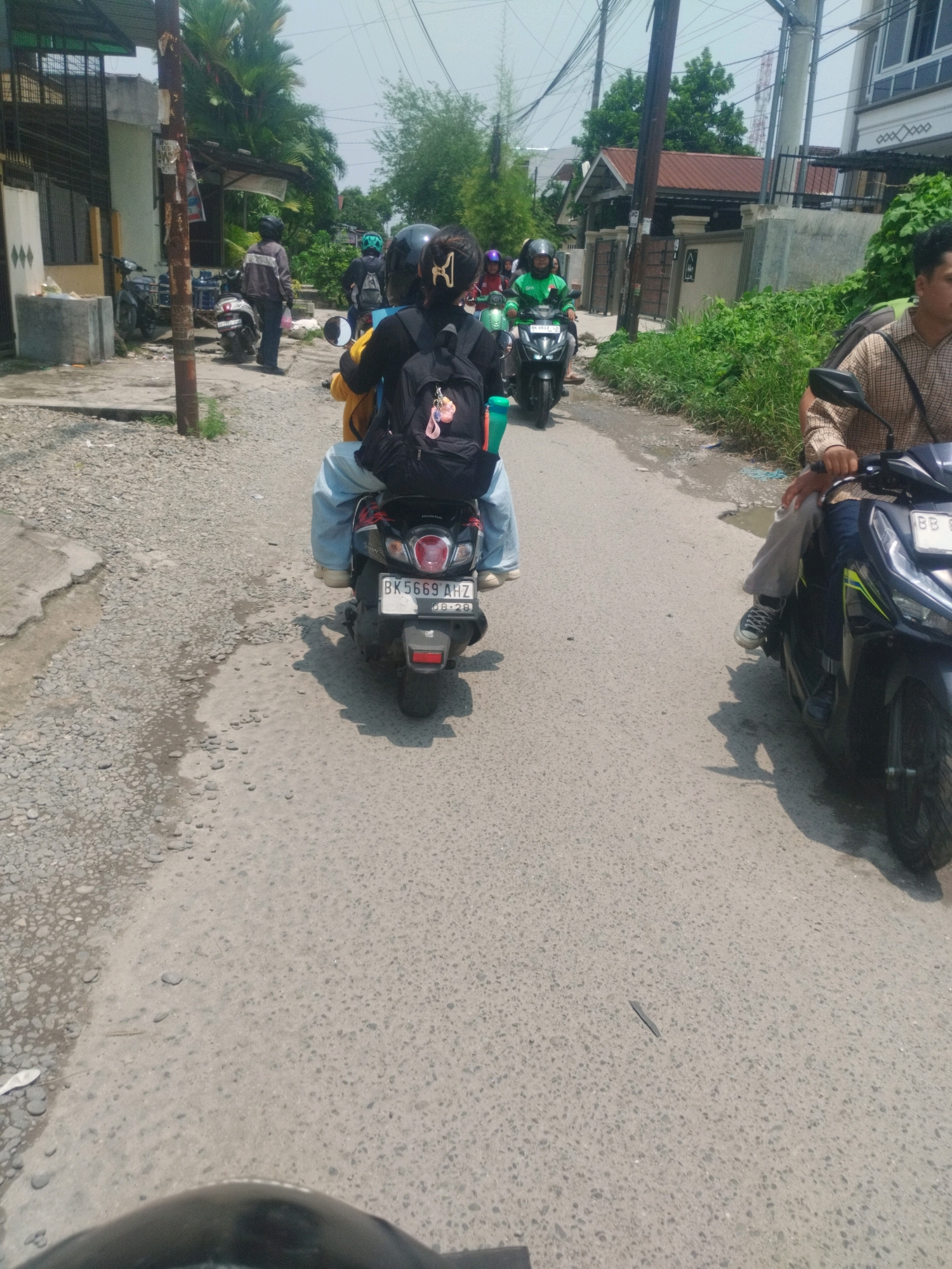 In a city street in Medan, Indonesia, three motorcycles are visible on a paved road. The closest motorcycle, viewed from the rear, has a license plate reading "BK 5669 AHZ". The rider is wearing a black backpack, and their legs are clad in light blue jeans and white sneakers. To the left of this motorcycle, another person is partially visible, also on a motorcycle. Further ahead, a person in a green jacket and helmet rides a motorcycle with a green "GoJek" logo on the side, and the license plate "BK 6513 UZ". Another motorcycle with a rider in a brown checkered shirt is seen on the right. The environment is characterized by buildings on either side of the road, some with houses and shops. Lush green vegetation lines the sides of the street, and utility poles with numerous wires are present overhead. The road surface is a mix of asphalt and gravel, with some water puddles visible. The sky is bright and overcast.