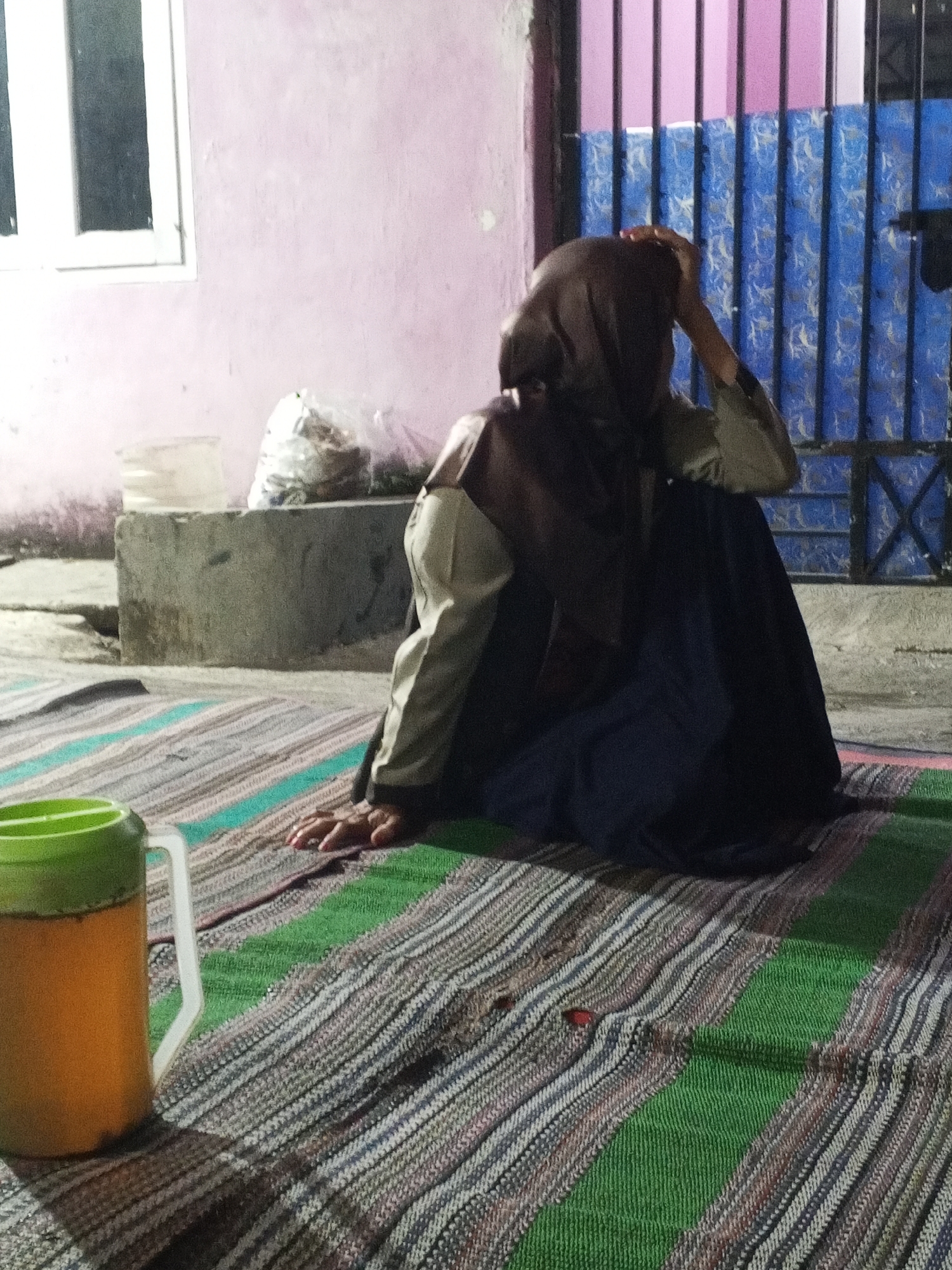 A person wearing a dark brown hijab and a light-colored shirt with dark sleeves is sitting on a striped mat on the ground. The person's left hand is resting on the mat in front of them, and their right hand is on their head. The person is facing away from the camera and to the left. In the foreground to the left, there is a large orange and green plastic jug with a white handle, casting a shadow. The background consists of a pink wall with a white-framed window on the left, and a blue metal gate with vertical bars on the right. A concrete block structure is visible behind the person. The scene appears to be outdoors at dusk or night, possibly in an urban or semi-urban setting in Ciruas, Indonesia.