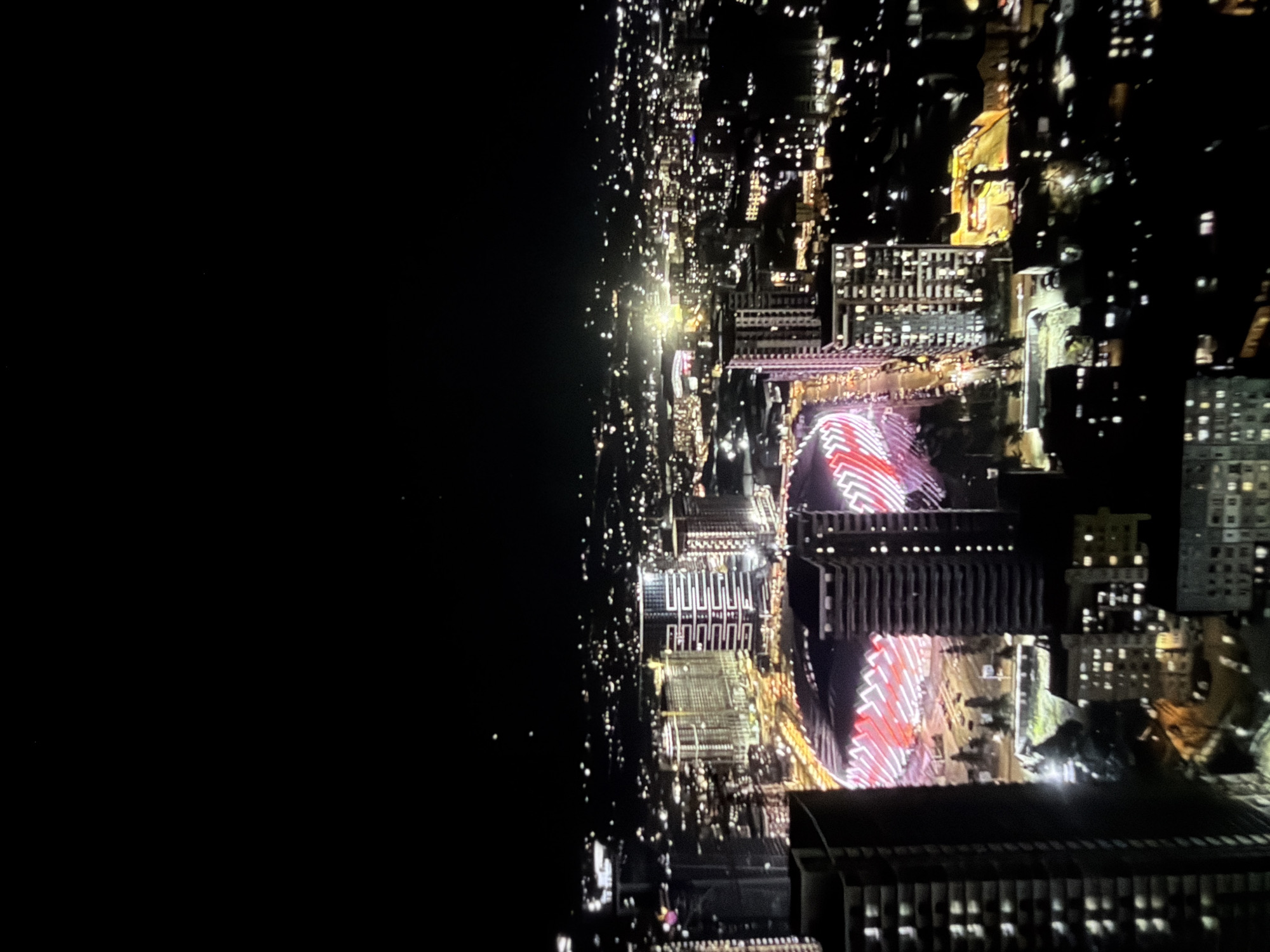 Here is a forensic description of the image:
**Scene:** Night aerial view of a cityscape.
**Location:** Unknown.
**Environment:** Dark sky to the left occupies a significant portion of the image. The right side shows densely packed city lights, illuminating buildings of varying heights.
**Objects:** Tall buildings are visible throughout the city, some with prominent red and white lighting patterns. Some buildings have visible lit windows.
**Light:** Primarily artificial, stemming from streetlights and building illumination.