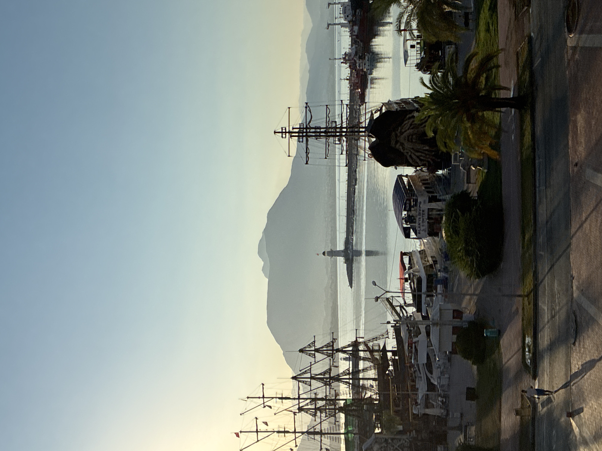 This is a vertical, medium-shot, full-color photograph taken from an elevated viewpoint, likely a hotel room or balcony, overlooking a harbor in Alanya, Turkey. The image captures the early morning or late afternoon light, with the sun positioned to the left, casting a warm glow and long shadows across the scene.
In the foreground, a paved walkway or promenade is visible, with a person walking from left to right, casting a long shadow. The walkway borders a grassy area with palm trees and other foliage. Beyond the walkway, a collection of boats and ships are docked. Prominently featured is a large, dark-hulled vessel with multiple masts, resembling a pirate ship. Other boats include smaller pleasure craft and what appear to be tour boats, one with a blue canopy and signage indicating "ALANYA DIVING CENTER".
The middle ground shows the calm harbor water, reflecting the light and the ships. In the distance, a pier extends into the water, with a lighthouse situated at its end. The lighthouse is white with a red-capped top. Beyond the harbor, a range of silhouetted mountains forms the backdrop, their peaks softened by the atmospheric haze. The sky is a gradient of pale blue, lighter near the horizon with the sun's influence, and deepening towards the top of the frame. The overall atmosphere is serene and picturesque, highlighting the coastal beauty of Alanya.