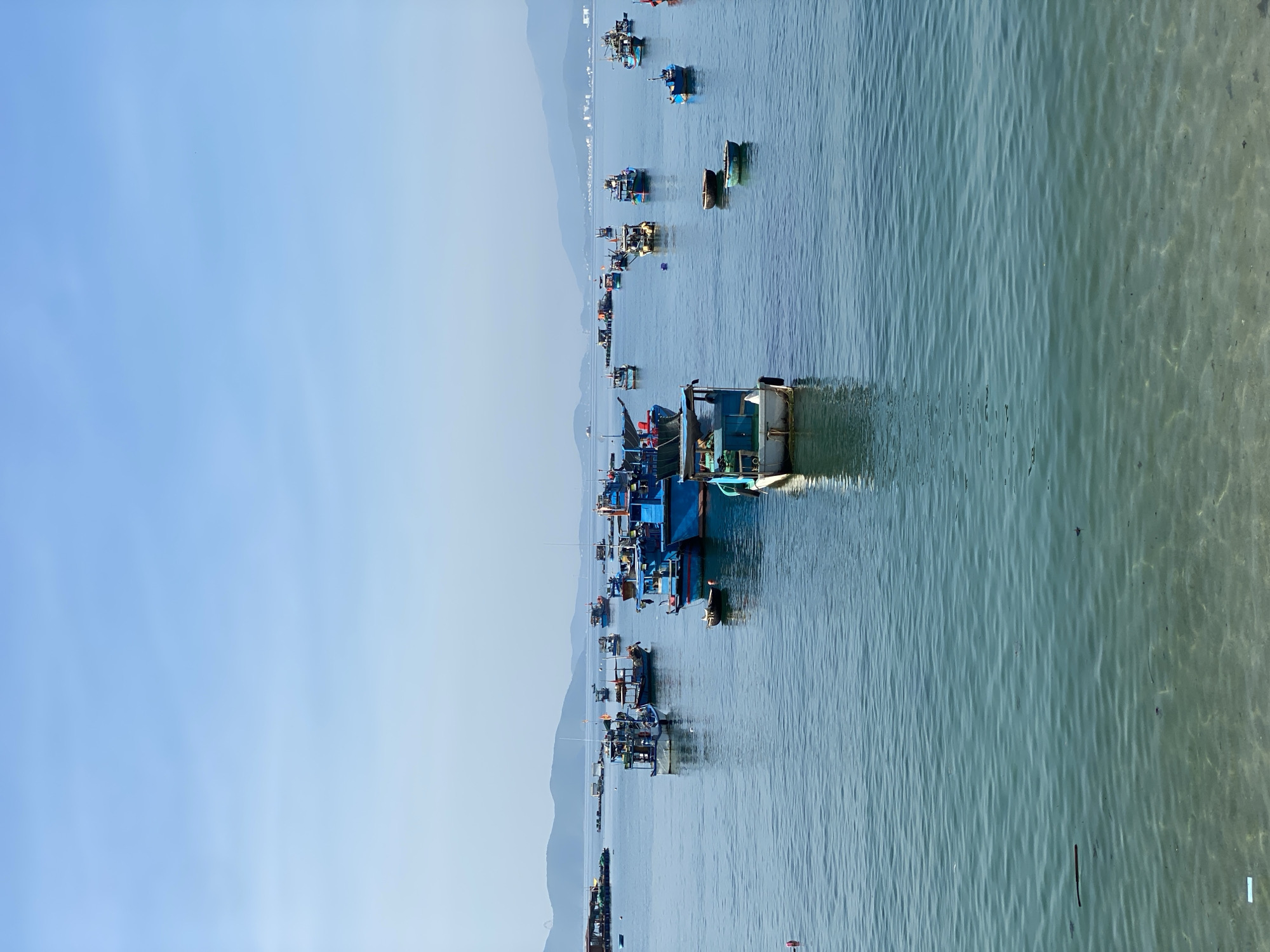This image depicts a bay filled with numerous fishing boats docked in shallow waters. The boats are of varying sizes and colors, with many appearing to be constructed of wood and painted in blues, whites, and greens. Several of the boats have multiple decks and structures, suggesting they are actively used for fishing. Some boats are moored together, while others are scattered across the bay.
In the background, a hazy coastline with mountains is visible, indicating a coastal or island setting. The water is calm and appears shallow near the shore, with a sandy or muddy bottom visible in places. The sky is a clear, pale blue, suggesting it is daytime, likely morning or afternoon, with good visibility. There are no people clearly visible in the image, and the boats appear to be either docked or anchored, implying a period of rest or preparation rather than active fishing at this moment.
The scene is peaceful and serene, with the stillness of the water reflecting the tranquil atmosphere. The overall impression is that of a working fishing harbor or a bay where fishing vessels are kept when not at sea. The location context of Bac Nha Trang Ward, Vietnam, further reinforces the image of a coastal community reliant on maritime activities. No visible text is present in the image.