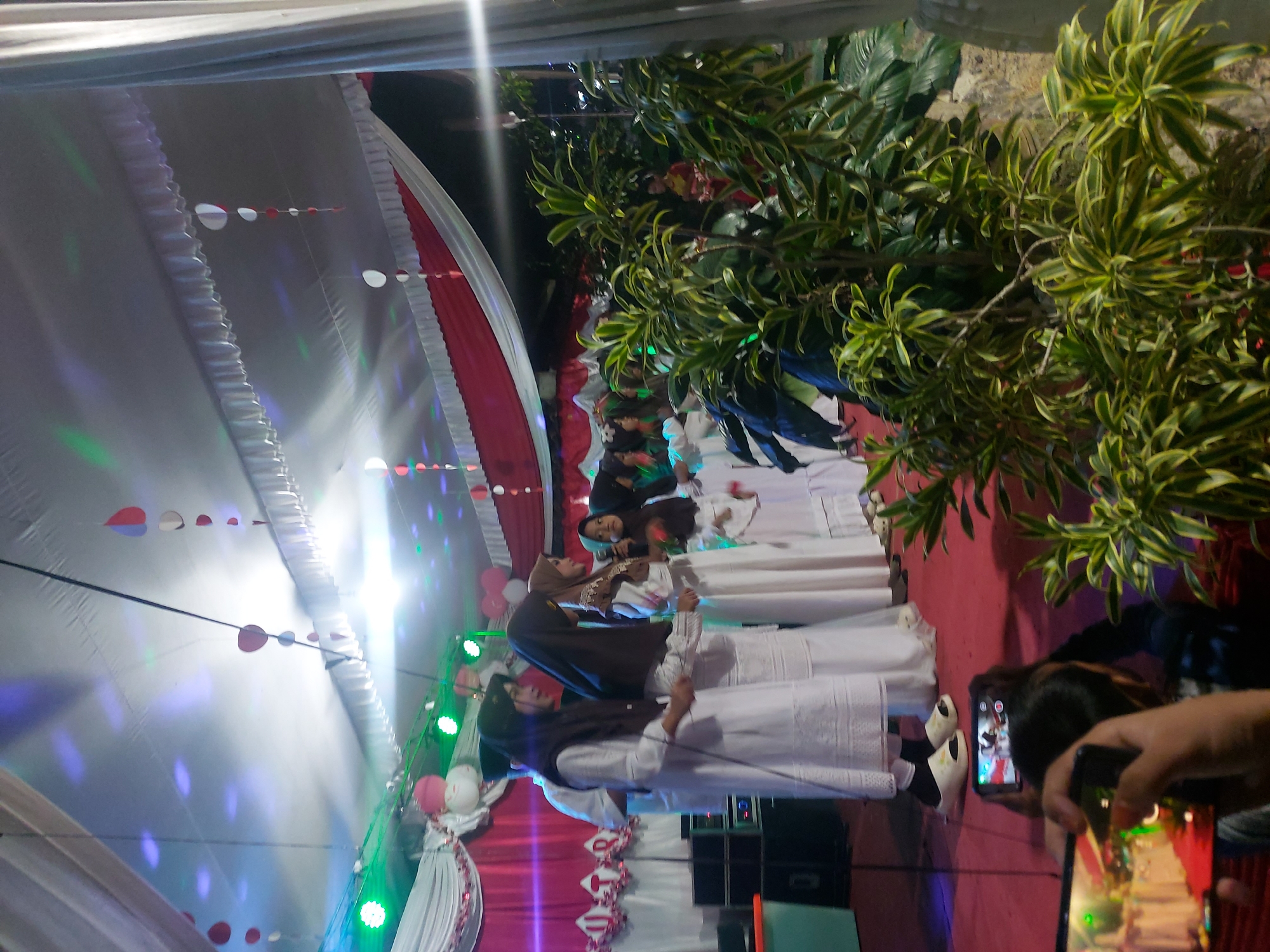 The image depicts an indoor event, possibly a school performance or ceremony, taking place under a tent adorned with white and red drapes, paper decorations, and colored lights. A group of young women wearing hijabs and white dresses stand on a red carpeted stage. In the foreground, two smartphones are being used to record the event. The background features green plants and some attendees. The lighting suggests it is evening or night. The event appears to be happening in Bandar Lampung, Indonesia.