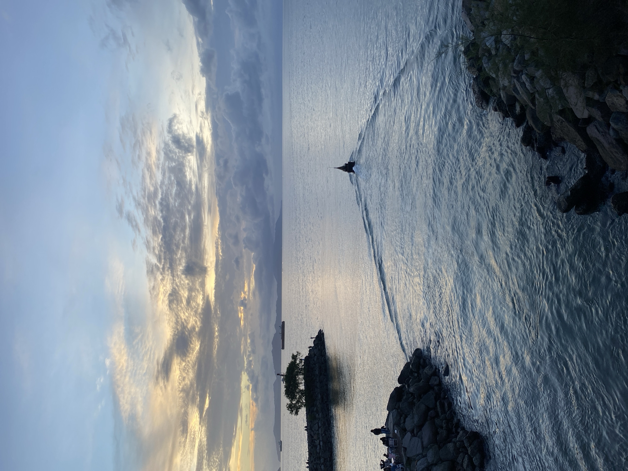 This image captures a serene seascape at dusk, likely taken from a coastal viewpoint in Banda Aceh, Indonesia, given the presence of a distinctive type of fishing boat common in the region. The sky is a canvas of soft blues and grays, with streaks of warm yellow and orange light peeking through dramatic, textured clouds, indicative of an impending sunset or afterglow.
The water is calm, reflecting the muted colors of the sky, and shows gentle ripples. A lone, dark-colored fishing boat, characterized by its pointed hull and raised stern, is moving away from the viewer, leaving a V-shaped wake on the water.
On the left side of the image, a rocky breakwater juts out into the sea. A small cluster of people are visible on this breakwater, some sitting on chairs, suggesting they are spectators or anglers enjoying the evening view. Further along the breakwater, a small island with a solitary tree stands as a focal point.
In the distance, a hazy coastline with rolling hills can be seen, and several large cargo ships are anchored or moving slowly on the horizon, indicating the area's maritime activity. The foreground is dominated by more rocky shorelines, with some sparse vegetation. The overall impression is one of tranquility and the end of a day by the sea.