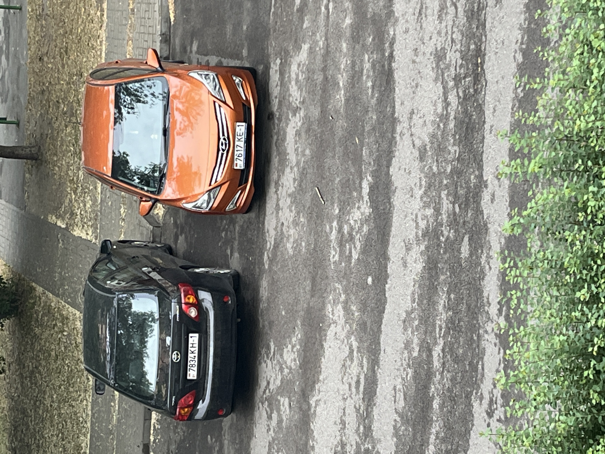 The image displays an overhead view of two parked cars on an asphalt surface, with a light-colored shoulder or path to their left and green foliage on the right. The image is rotated 90 degrees clockwise, placing the cars vertically.
The car positioned closer to the viewer is an orange or copper-colored Hyundai, identifiable by its front emblem and grille design, likely an Elantra model. Its front license plate reads "7617 KE-1" and features a small Belarusian flag icon.
Further down the asphalt, behind the orange car, is a black Toyota Corolla, with "TOYOTA" and "COROLLA" badging visible on its rear. Its rear license plate displays "7834 KH-1," also featuring the Belarusian flag icon.
The scene is set outdoors during daytime, under what appears to be an overcast or partly cloudy sky, as evidenced by the diffused light and lack of strong, sharp shadows. The asphalt shows variations in texture and some damp-looking streaks. No people are visible, and the cars are stationary, suggesting they are parked along a street or in a parking area in Belarus.