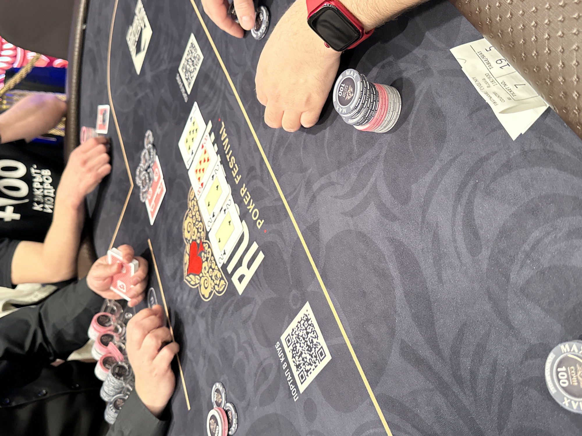 A poker game is in progress on a gray felt table with the words "RU POKER FESTIVAL". Several players are visible, with chips stacked in front of them. The table has QR codes and designs on it. The community cards on the table are visible, and a ticket with "FIRSTNAME: EVGENIY, COUNTRY: Russia, TABLE/SEAT: 19 5" is present. Some poker chips have the words "MAX 100 COЧИ". The location is most likely in Estosadok, Russia, given the Cyrillic text on the chips and the player's country on the ticket.