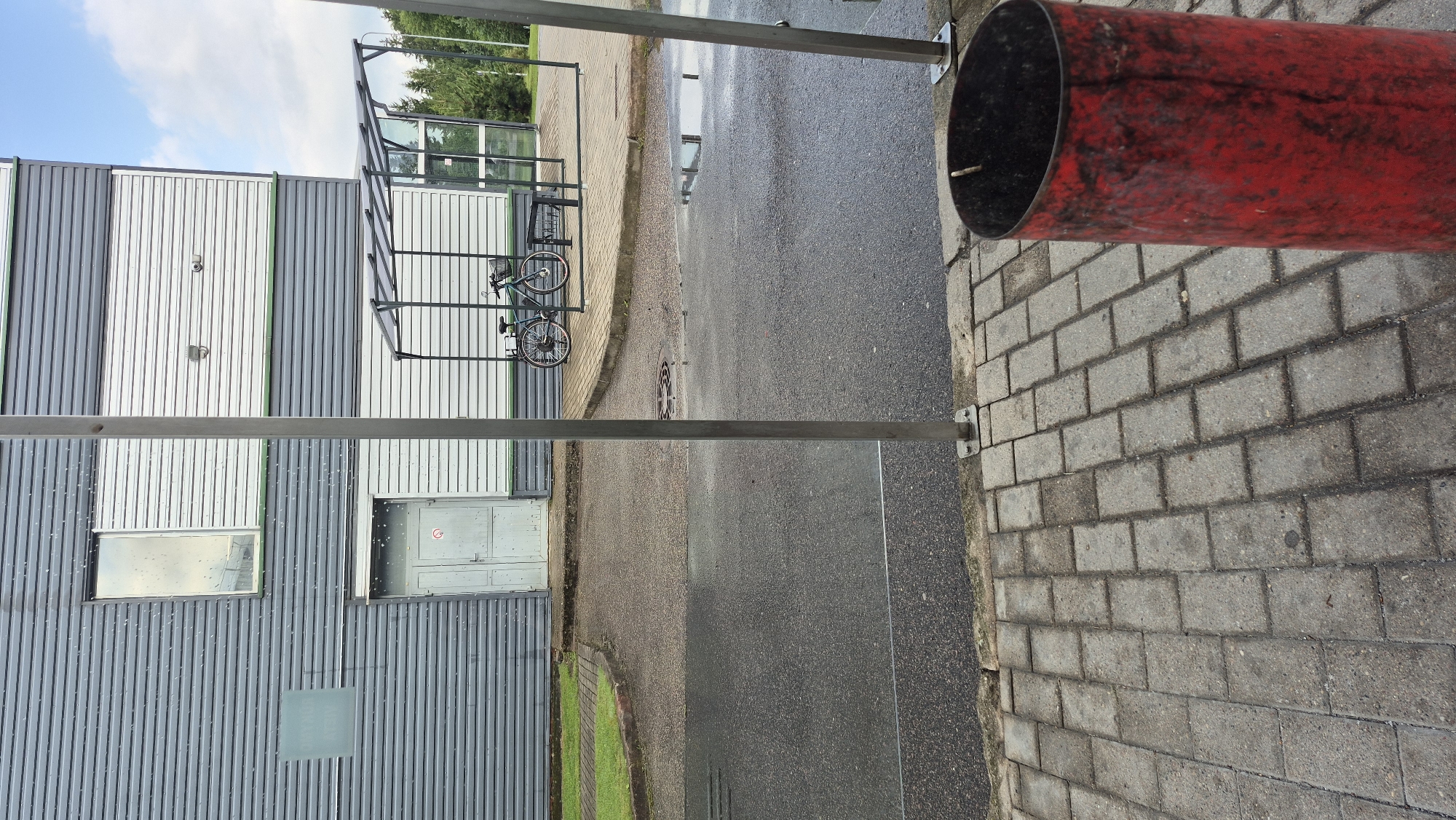 The image depicts an outdoor scene, likely in the city of Pabradė, Lithuania. In the foreground, a red cylindrical trash can is positioned on a paved walkway made of rectangular bricks. Beyond the walkway, a wet asphalt road stretches across the frame, with several puddles reflecting the sky and surrounding structures. A metal pole stands vertically in the mid-ground, positioned on the edge of the walkway and road.
In the background, a two-story building with a modern, industrial facade is visible. The building's exterior is clad in corrugated metal siding, alternating between grey and white panels. One prominent feature is a large, rectangular window on the upper level, reflecting the cloudy sky. A closed grey double door is situated on the lower level of the building, with a circular "no smoking" symbol on it.
To the right of the building, a small covered area with a transparent roof and glass panels serves as a bike shelter. A single bicycle, predominantly dark green with a basket on the front, is parked inside the shelter. Further to the right, trees and foliage indicate a natural environment bordering the developed area. The sky is partly cloudy with blue patches. The overall impression is of an urban or industrial setting during or after rainfall, given the wet surfaces and puddles.