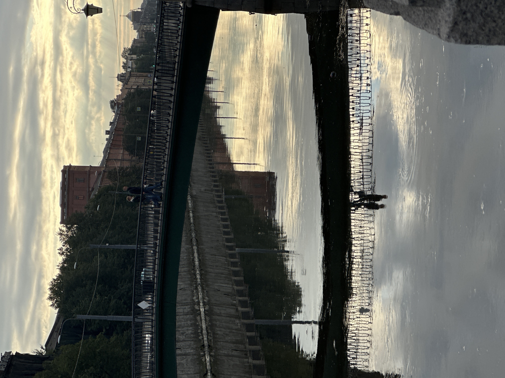 The image shows a bridge over a canal in Saint Petersburg, Russia. The sky is overcast with some rays of sunlight breaking through. There are buildings and trees on either side of the canal, and the reflection of the bridge and buildings can be seen in the water. Two people are walking across the bridge. The overall mood of the image is peaceful and serene.