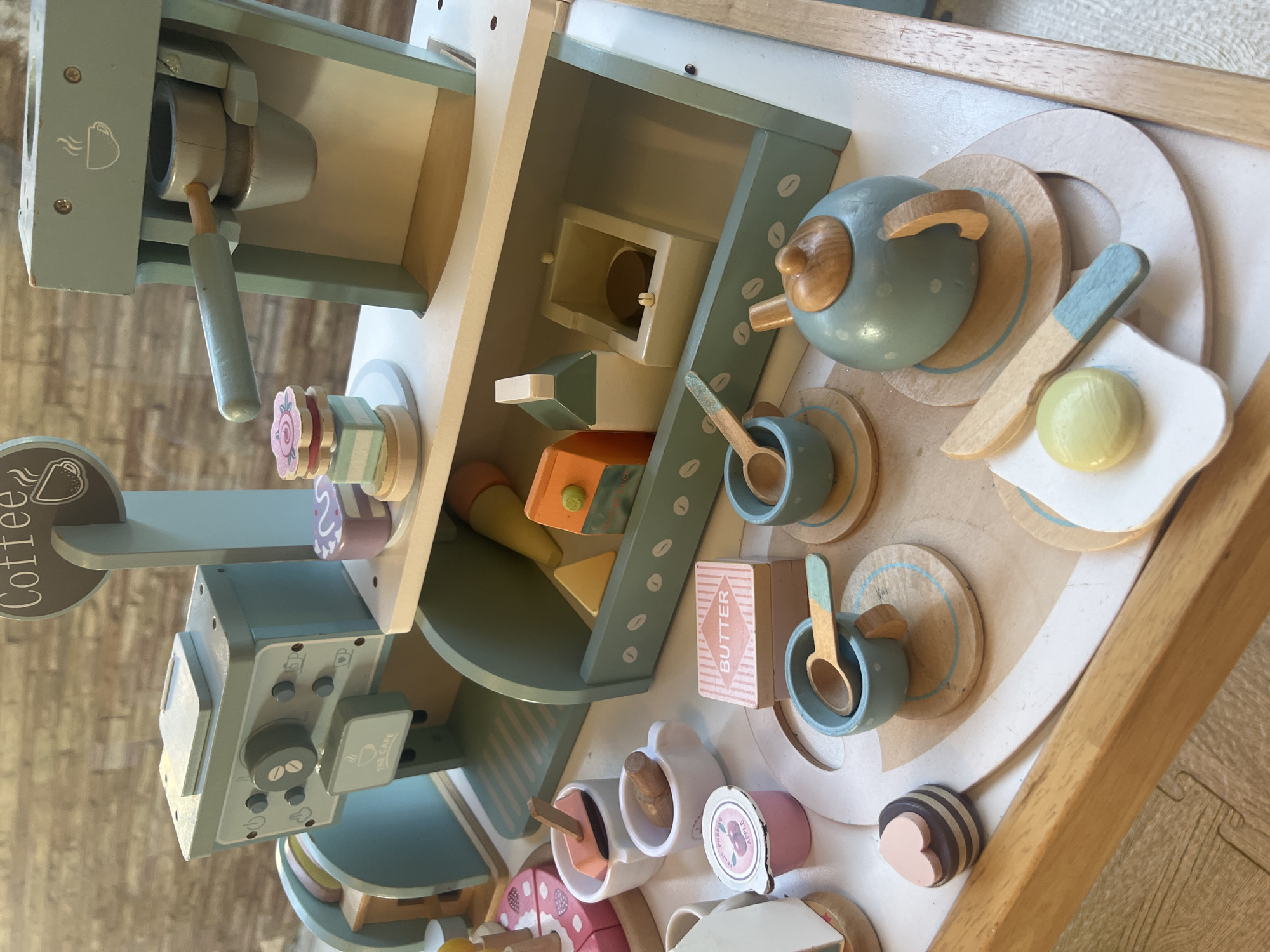 A wooden toy coffee shop playset is displayed on a wooden surface. The playset includes a coffee machine, cups, a teapot, a butter box, a cake, a carton of juice, and other accessories. The backdrop appears to be a textured wall. The location of the image is Ko Samui District, Thailand.