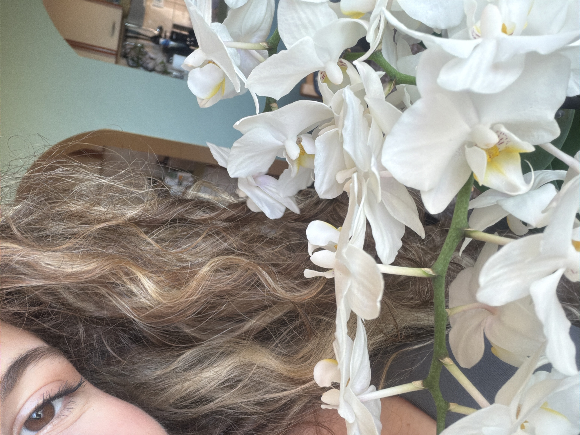 The media file is a close-up, high-angle shot of a person's face and hair, partially obscured by a cluster of white orchids. The person, who appears to be female, is on the left side of the frame. Only her left eye, eyebrow, and a portion of her cheek are visible. Her eye is brown, with winged eyeliner and mascara. Her hair is long, wavy, and a blend of blonde and light brown tones. The orchids are delicate and pure white, with subtle yellow and reddish markings in their centers. They are arranged on a green stem, with some blossoms hanging over the person's hair. The background is a soft, pale blue, with a reflection of what appears to be an indoor setting with furniture and a mirror in the upper left corner. The overall impression is one of tranquility and natural beauty, possibly suggesting a relaxed moment. No text is visible in the image. The location context is Lyubimets, Bulgaria, but this is not discernible from the image itself.