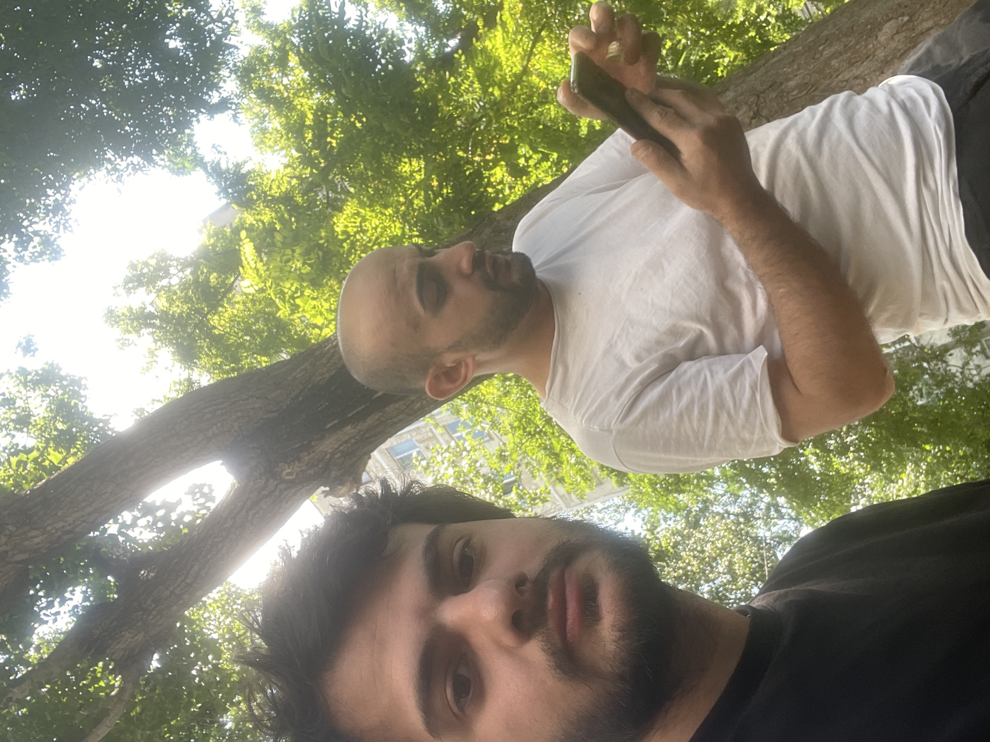 Two men are in an outdoor urban setting, likely Tbilisi, Georgia, with lush green foliage and tree branches in the background. The man in the foreground, on the left, is captured in a close-up, his face filling the frame. He has dark, tousled hair and a beard, and is looking directly at the camera with a neutral expression. He is wearing a black t-shirt. To his right, partially obscured by the foreground man, is another man with a shaved head and a light beard. He is wearing a white t-shirt and is looking down at a smartphone he holds in his hands, with his right hand's index finger extended as if interacting with the screen. A ring is visible on his right ring finger. The bright sunlight filters through the leaves, creating a dappled effect and partially overexposing the scene, particularly the sky and upper foliage. A building facade with windows can be seen faintly through the leaves in the mid-ground. The presence of trees and buildings suggests a park or a tree-lined street within the city.