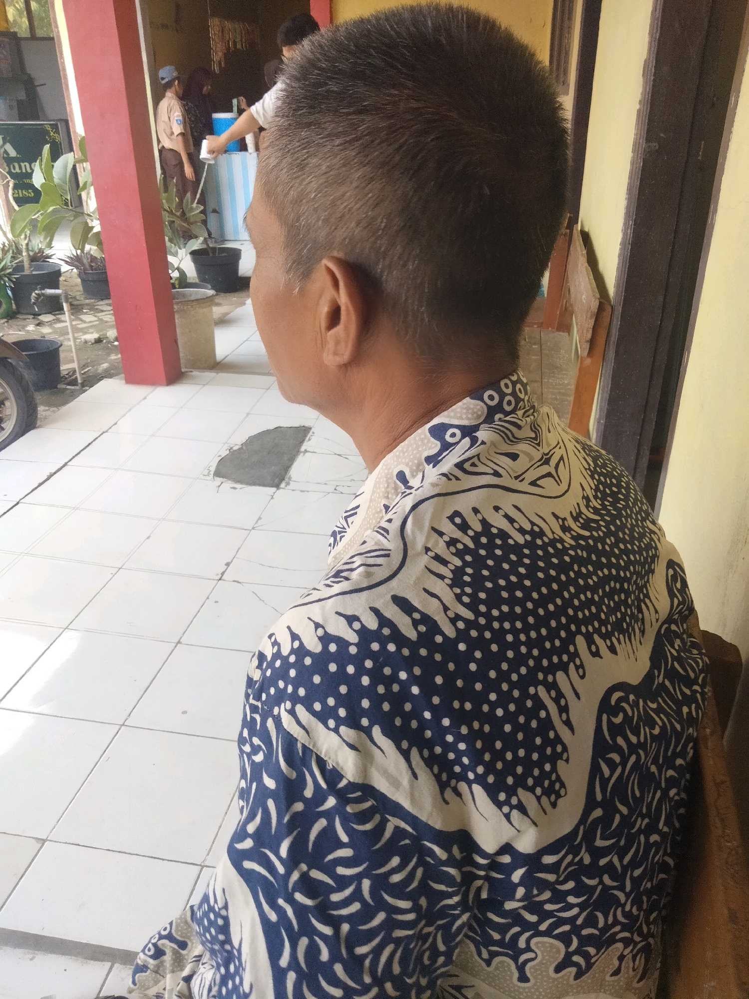 In the city of Suka Sari, Indonesia, a medium shot captures a person from behind, seated and facing away from the camera. They are wearing a white shirt with a distinctive blue and white patterned design, possibly batik. The person has short, graying hair. The setting appears to be outdoors, with a tiled floor and a red-painted pillar visible on the left. In the background, other individuals are present, including one wearing a brown uniform and a blue cap, possibly a student or scout, who is interacting with a water dispenser. Several potted plants and a motorcycle wheel are also visible. The lighting suggests it is daytime.