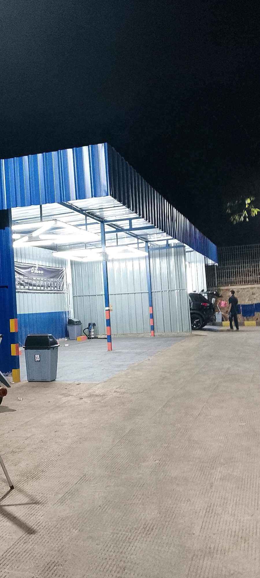 The image shows a car wash facility at night in Gunung Putri, Indonesia. The facility has a blue and white corrugated metal roof and walls, with fluorescent lighting illuminating the interior. A dark-colored SUV is parked inside the wash bay, and a person in dark clothing is standing near it. In the foreground, there is a gray trash bin with a black lid, and a vacuum cleaner is visible behind it. The ground is a concrete surface with a grooved texture. A dark sky is visible above the structure, with some distant foliage on the right side.