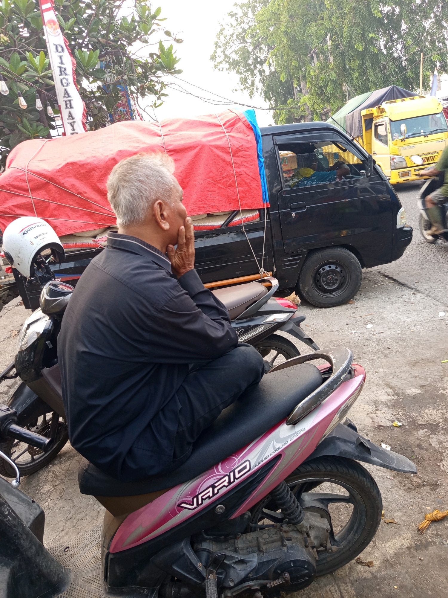In Karawang, Indonesia, an elderly man with gray hair sits on a pink and black scooter, his face obscured as he rests his chin on his hand. He wears a dark long-sleeved shirt and dark pants. The scooter is parked on a paved surface, with its front wheel visible to the left and its rear wheel and shock absorber clearly shown on the right. Next to the scooter, another motorcycle with a white helmet on its handlebars is parked. In the background, a black pickup truck is parked, its bed covered with a red tarp. A yellow truck is also visible further back. Trees and a flag bearing the Indonesian text "DIRGAHAYU" are present in the upper left portion of the image. The scene appears to be a roadside in Karawang.