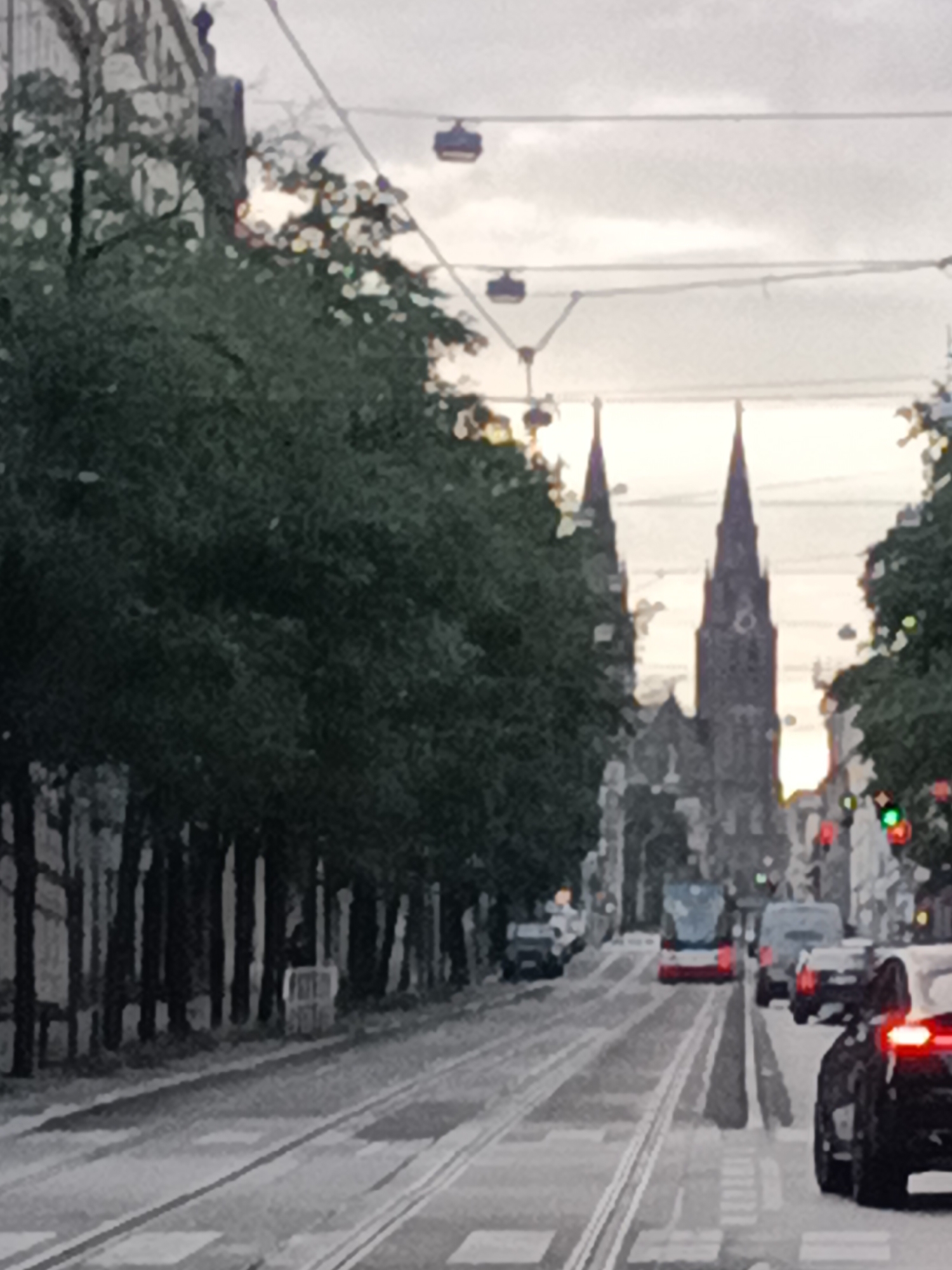 The image depicts a street scene in Prague, Czechia, likely during overcast conditions. The perspective is from street level, looking down a road with tram tracks. On the left side of the street, dense foliage from trees lines the road, with some buildings visible behind them. In the distance, two prominent church spires rise above the tree line. Several vehicles are present on the road, including a red and white tram in the middle ground and several cars. The sky above is a bright, overcast white. Tramway wires are suspended overhead, and a few cable car-like structures are also visible, suspended from the wires. The scene suggests an urban environment with public transportation infrastructure and historical architecture.
