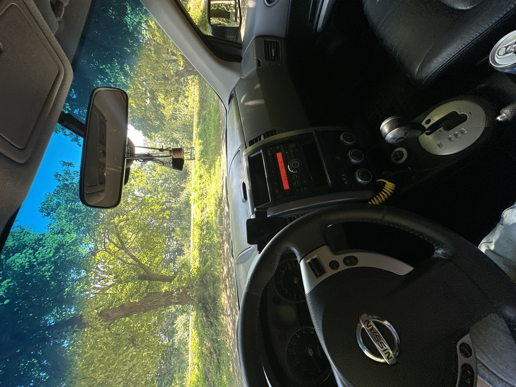 The image captures the interior of a Nissan vehicle, seen from the driver's perspective. The dashboard and steering wheel are prominent, with the Nissan logo visible on the steering wheel. The car's radio display shows "FM2 90.8". A gear shifter is visible, along with part of the passenger seat. Outside the car, through the windshield and side windows, lush green trees and foliage are seen under a bright sky, suggesting an outdoor, possibly rural, setting. The sunlight filtering through the trees creates dappled patterns on the dashboard. A rear-view mirror hangs from the windshield, with an air freshener attached. The scene appears to be static, with no visible people or actions taking place. The location is described as Shatava, Ukraine.