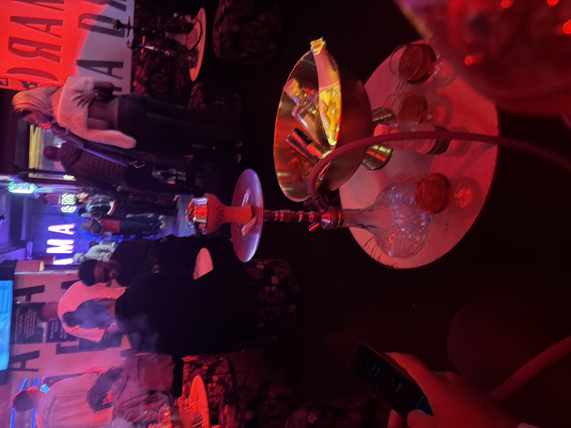 The image shows a dimly lit indoor scene, likely a bar or lounge, with a hookah prominently displayed on a circular table. Multiple glasses, possibly containing alcoholic beverages, surround the hookah. Several people are visible in the background, some standing and some seated. A person is holding a phone, likely operating the hookah. The background includes signage with partially legible words like "IMA" and "MARGARITA." Based on this signage, the location is the MARGARITA bar and lounge in Cape Town, South Africa. There is activity within the bar, implying social interaction and possibly consumption of hookah and drinks.