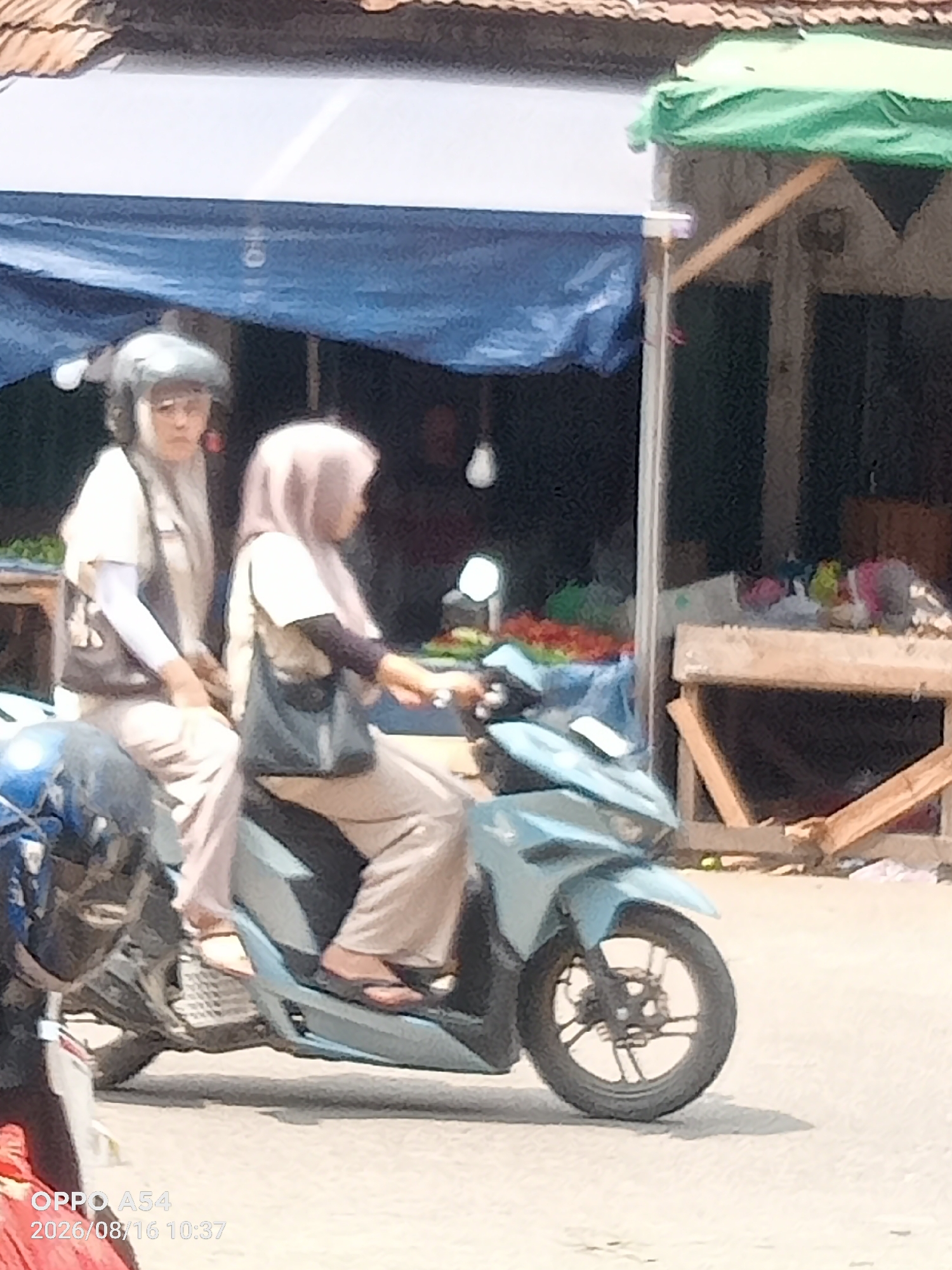 The image captures a scene in the city of Jambi, Indonesia, featuring two individuals on a light blue scooter. The person in the front, likely the driver, wears a light-colored hijab and a matching long-sleeved shirt. The person behind, possibly a passenger, is also wearing a light-colored hijab and a cream-colored long-sleeved top, with white arm coverings visible. Both individuals are wearing light-colored pants. The scooter has a dark-colored seat, and the front wheel is clearly visible. The background suggests an outdoor market setting with a blue tarp overhead, wooden structures, and various items for sale, including what appear to be colorful bags or fabrics. A wooden stall with diagonal supports is prominent on the right side of the image, with a green tarp draped over part of it. The ground is paved with a textured surface. The lighting indicates it is daytime.