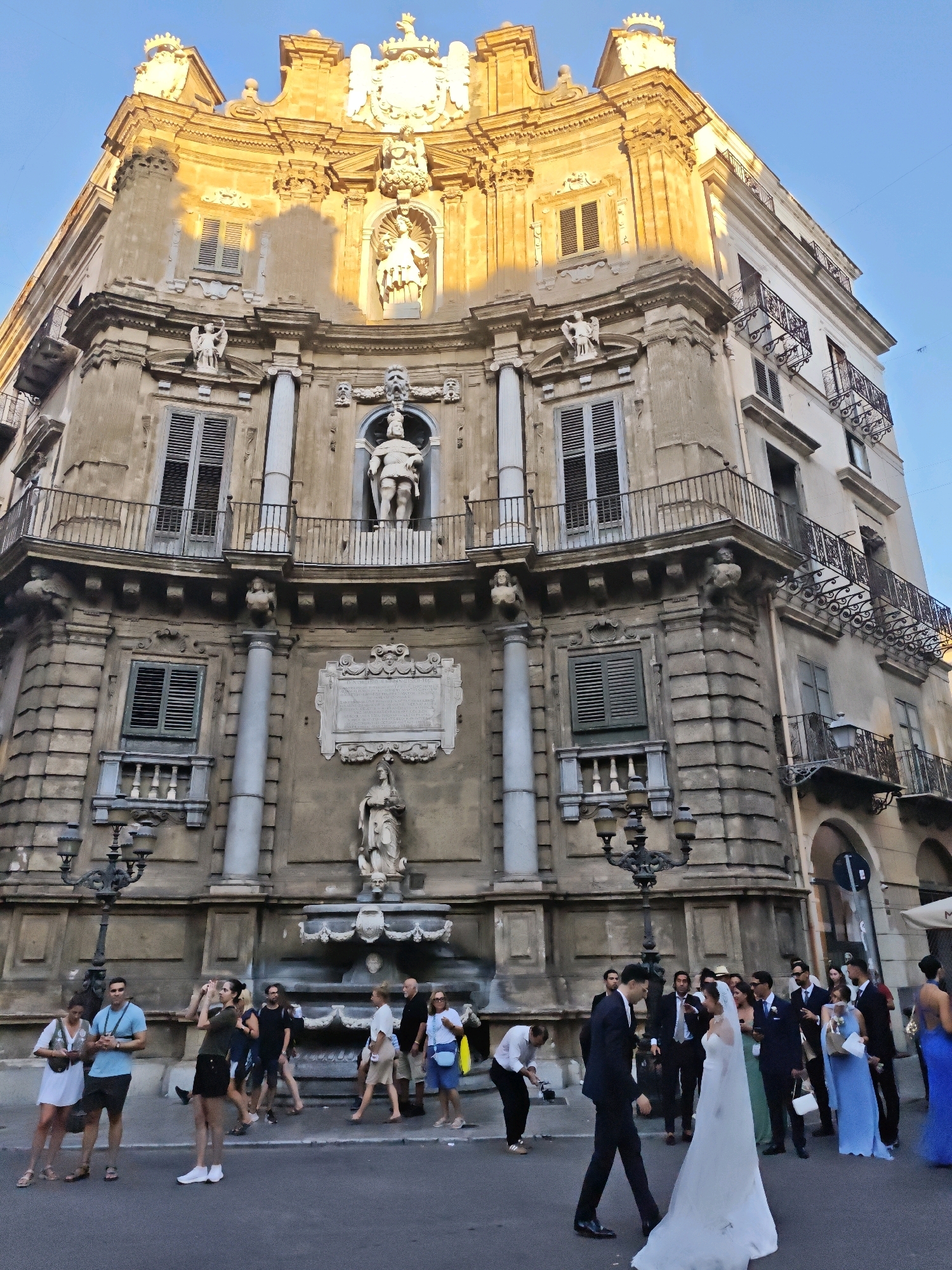 The image shows the Quattro Canti, a Baroque square in Palermo, Italy, at what appears to be a wedding reception. The Quattro Canti is characterized by its four ornate buildings at the intersection of Via Maqueda and Corso Vittorio Emanuele. In the foreground, a bride in a white wedding dress and a groom in a dark suit walk towards each other. They are surrounded by wedding guests, some of whom are dressed in formal attire, while others are in more casual clothing. A woman in a white dress and a man in a blue t-shirt are standing to the left, seemingly observing the wedding party. A photographer with a camera is visible to the left, capturing the moment. The architecture of the Quattro Canti, with its statues and decorative elements, is prominently featured in the background, bathed in the warm light of the sun. The sky is clear and blue.
