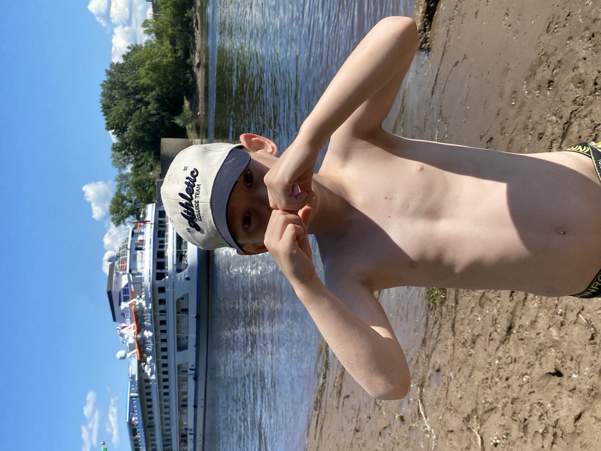 The image shows a young boy in a bathing suit standing on a sandy shore next to a body of water. He is wearing a baseball cap that says "Athletic College Team" and has the number "91" on it. He is holding his hands up to his face and looking at the camera with a playful expression. Behind him, a large white boat with multiple decks is docked on the water, and trees line the opposite shore. The sky is blue with some white clouds, suggesting it is a sunny day. The sandy shore is wet in places, indicating that the water level is high or has recently receded. The overall impression is a casual summer day spent by the water.