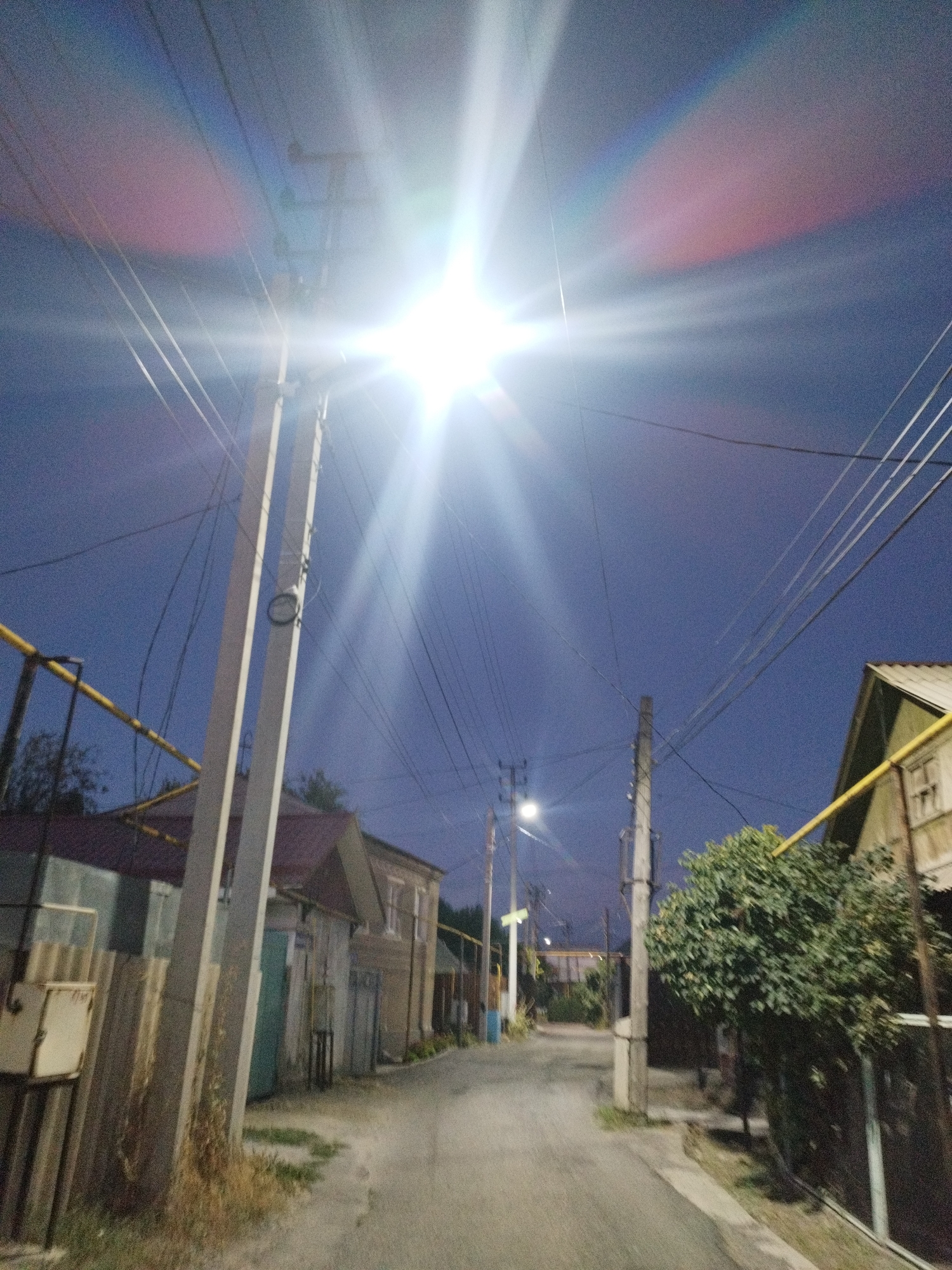The media file depicts a dimly lit street in a residential area, likely at dusk or dawn, given the clear blue sky and the bright streetlamp illuminating the scene. The setting appears to be Shymkent, Kazakhstan. A prominent streetlamp, the source of the intense light and lens flare, is mounted on a utility pole positioned near the center of the frame. Power lines crisscross the sky, adding to the urban feel of the scene.
The street itself is paved with asphalt and curves gently into the distance. On either side of the street are houses with visible architectural details such as sloping roofs and windows. Some of the houses appear to be older, with visible wooden or corrugated metal siding. A bushy green tree is visible on the right side of the street, its foliage illuminated by the streetlamp. Yellow gas pipes are also visible running along the exterior of some of the buildings.
There are no people visible in the frame. The overall atmosphere is quiet and somewhat still, with the primary visual focus being the strong artificial light against the darkening sky. The presence of utility poles, power lines, and gas pipes suggests a functional, lived-in neighborhood. The lens flare from the streetlamp creates a dramatic effect, with beams of light radiating outwards and a soft pink and blue hue appearing in the sky. No text is visible in the image.
