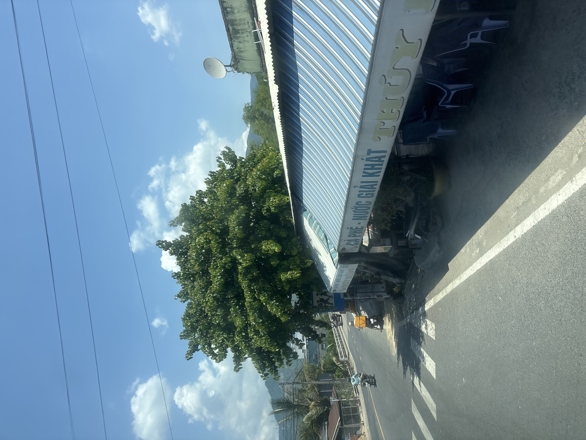 The image depicts a roadside scene in a tropical or subtropical environment. A paved road runs from left to right across the frame, with a pedestrian crossing marked by white stripes in the foreground. Two motorcycles are visible on the road; one in the left lane appears to be moving away from the viewer, while another in the right lane is stationary.
On the right side of the road, a building with a corrugated metal awning extends over a seating area. The awning is striped with shades of blue and white, and a sign in Vietnamese reads "CÀ PHÊ - NƯỚC GIẢI KHÁT THUÝ," indicating it is a coffee and soft drink establishment. Blue plastic chairs are arranged under the awning.
A large, leafy green tree dominates the middle ground, casting shadows on the road and the building. In the background, there are more trees, some structures, a bridge, and a body of water with distant hills. The sky is clear blue with scattered white clouds. A satellite dish is visible on the roof of the building. The overall impression is a casual, open-air establishment along a road in a developing area.