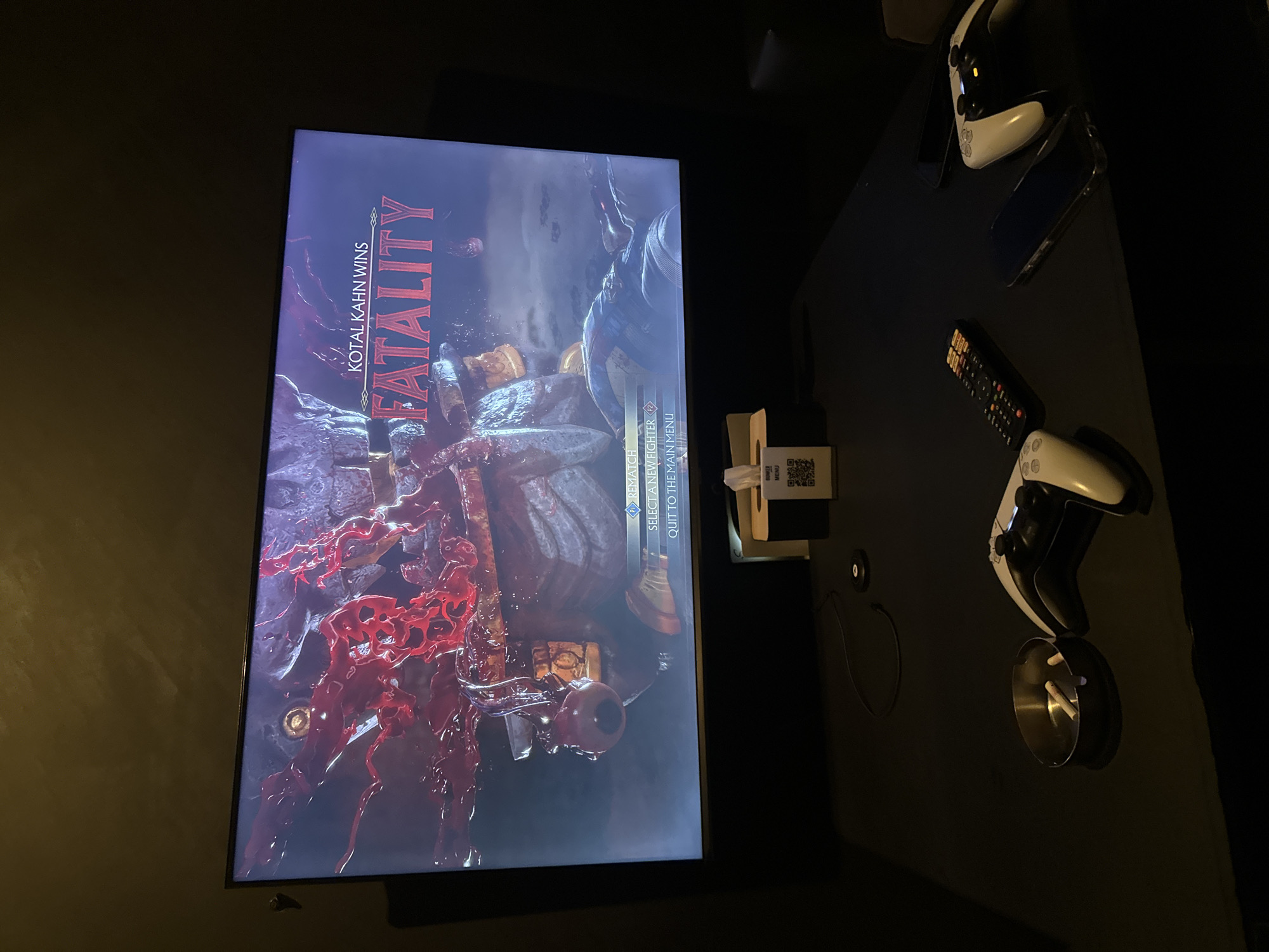 The image shows a television screen displaying a "FATALITY" message after Kotal Kahn wins in Mortal Kombat. The game screen shows blood and gore. The television is situated on a black surface. To the right of the screen are two PlayStation 5 controllers, two iPhones, and a TV remote. An ashtray with cigarette butts is also visible. A tissue box with a QR code on it is nearby. The location is unknown, and there's no indication that it is in Yerevan, Armenia. There are no people visible in the image.