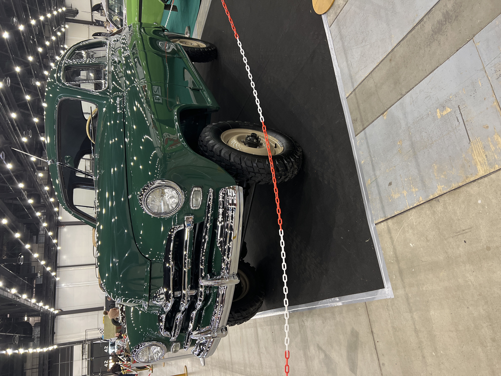 The image shows a green vintage car on display at a show, likely indoors. The car has a classic design with chrome accents and large tires. A red and white chain serves as a barrier around the display area. The location appears to be a large event space with overhead lighting and concrete flooring. The city is Unknown, Unknown.