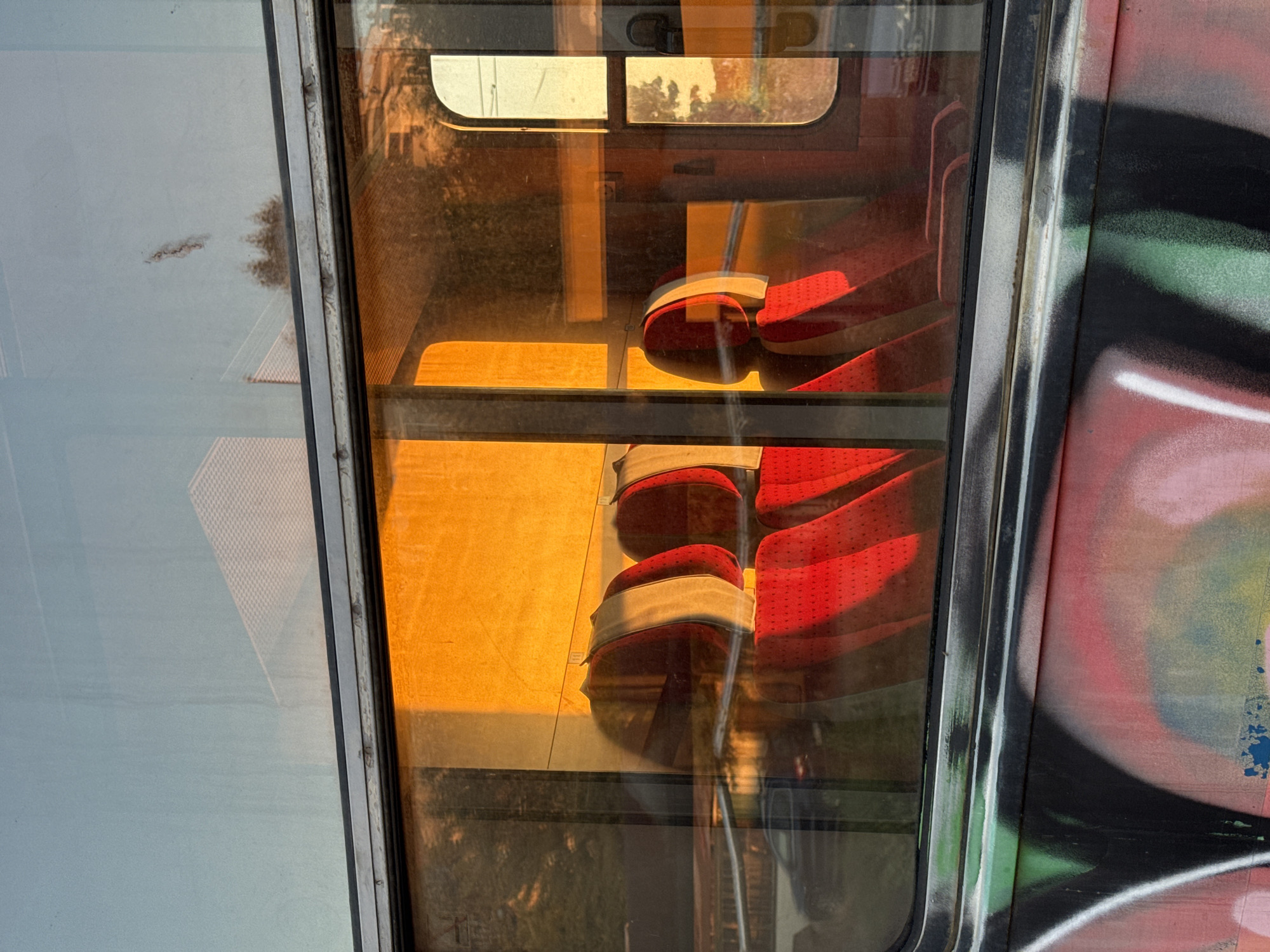 The image shows the interior of a train carriage through a window. The seats are upholstered in red fabric with white headrests. Sunlight is streaming through the window, casting a warm, orange glow on the interior. Reflections on the window include trees and buildings outside. The exterior of the train is covered in graffiti. The scene is likely located in Burgas, Bulgaria, given the context provided. There are no people visible in the image.