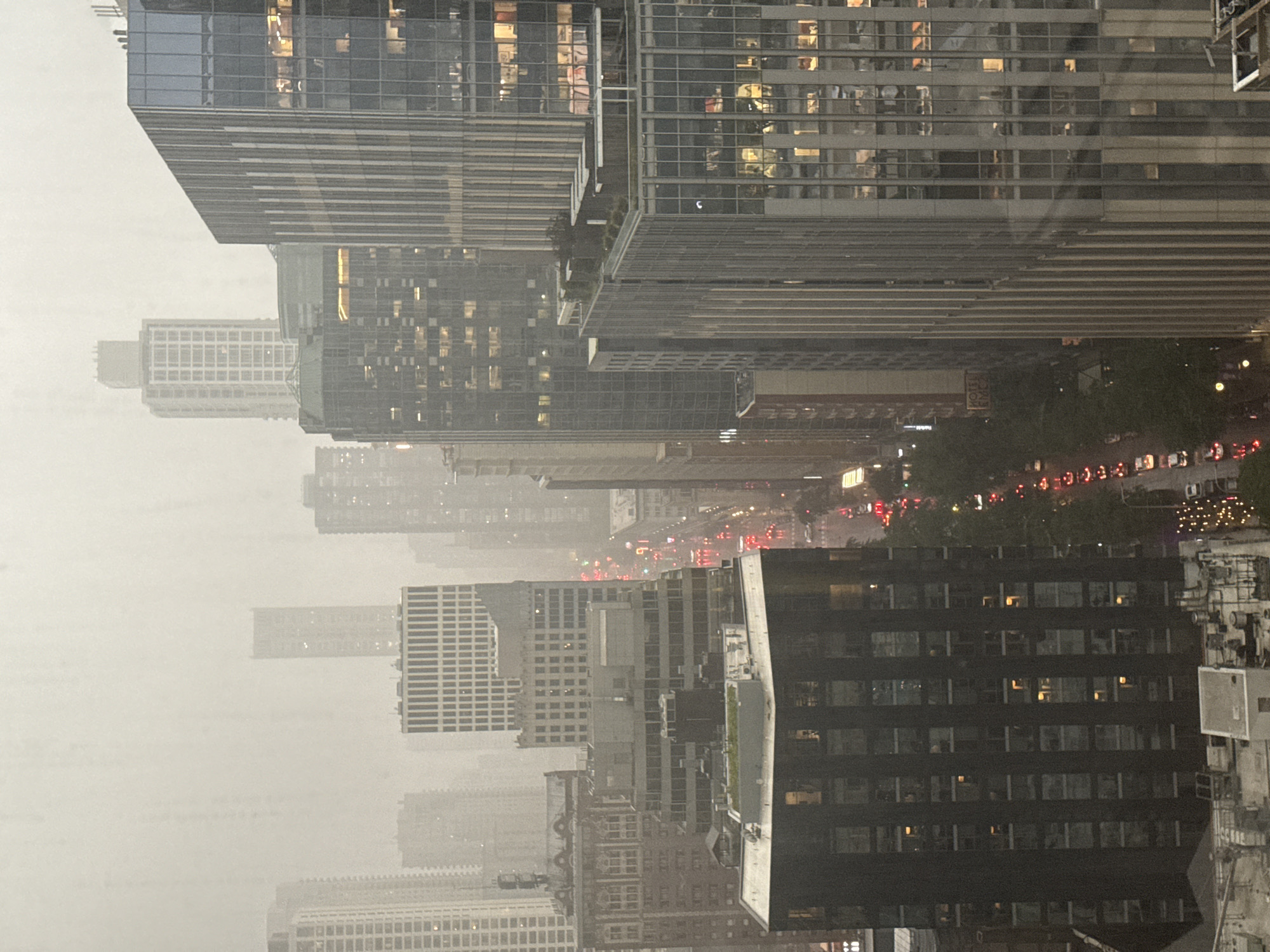 The image captures a hazy, overcast view of a dense urban landscape, likely Chicago, given the architectural style and the density of skyscrapers. The scene is dominated by tall buildings of various designs, from modern glass-fronted structures to older, more ornate buildings. A street level view is visible looking down a canyon of buildings, with a traffic stream indicated by rows of red taillights and white headlights, suggesting vehicles are moving in both directions. The weather appears to be inclement, with a pervasive fog or mist obscuring distant structures and softening the light, giving the scene a muted, almost ethereal quality. Some windows in the buildings are illuminated, indicating occupancy, while others are dark. The overall impression is one of a bustling city enduring a heavy fog or rain.