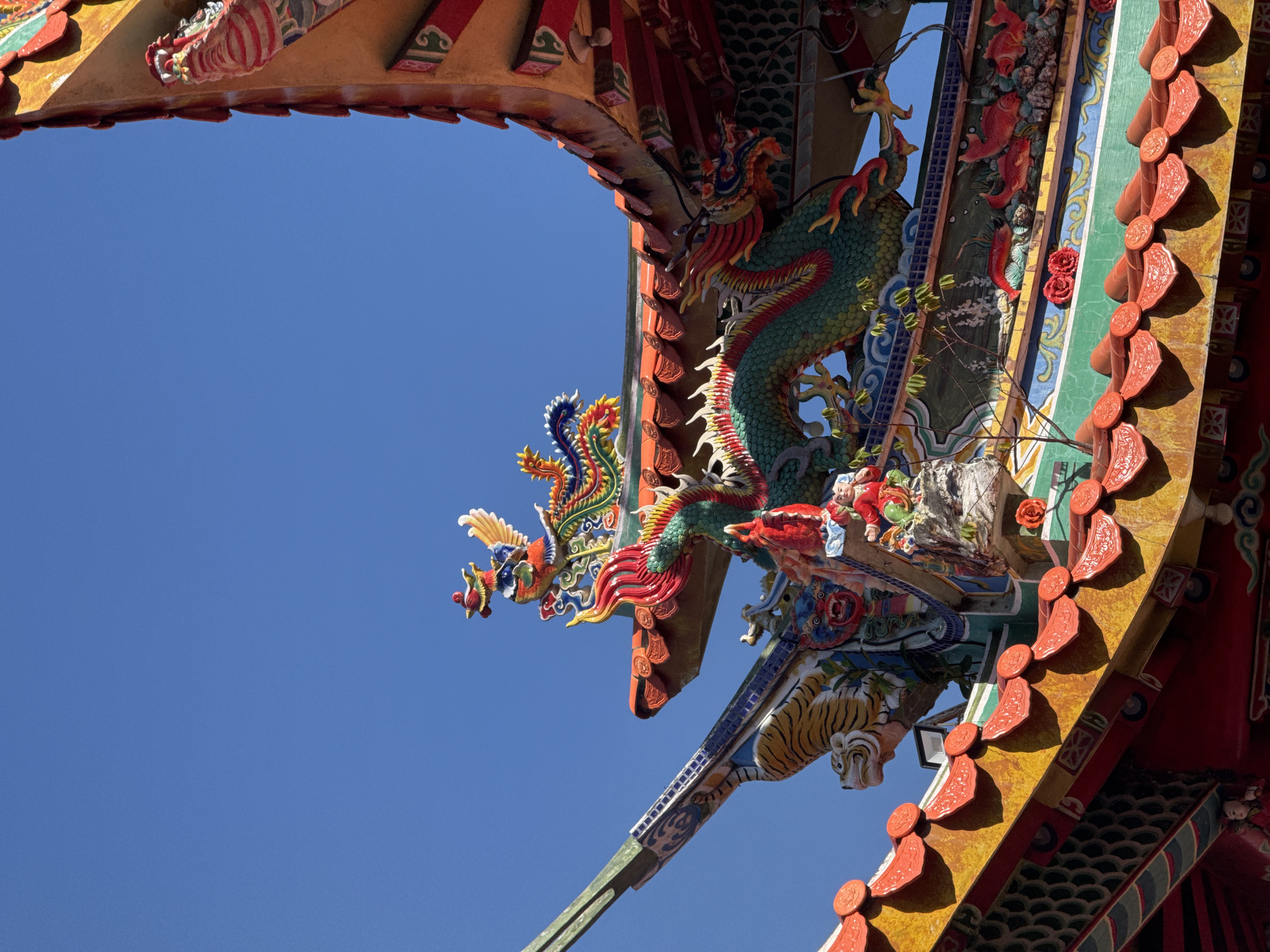 The image shows a portion of an elaborately decorated structure against a clear blue sky. The structure features intricate carvings and vibrant colors. Notable details include a multi-colored dragon figure with scales in green, red, and other colors, architectural details such as a tiger head, and decorative elements in shades of red, yellow, green, and blue. Based on the architectural style and the presence of dragon imagery, this is likely a religious or cultural structure found in Denpasar, Indonesia, although the specific location cannot be definitively determined from the image alone.