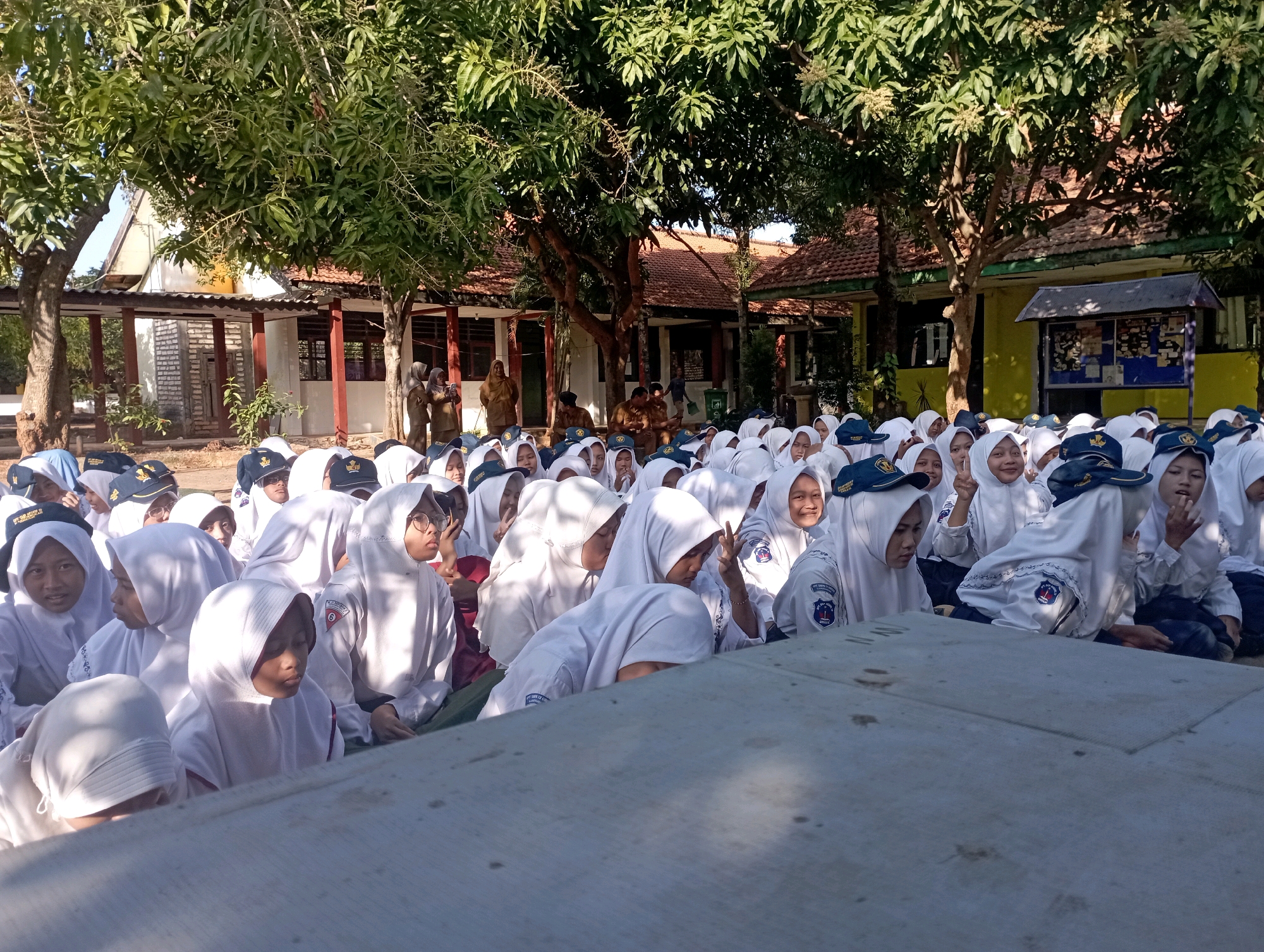 In the city of Banyuurip, Indonesia, a group of students wearing white uniforms and hijabs are gathered outdoors, likely on a school campus. The students are seated on the ground, facing towards the viewer. Many of them are looking at the camera, with some smiling or making gestures. Behind the students, a building with a red tiled roof and open-air corridors is visible, suggesting an educational institution. Large trees with green foliage provide shade over the scene. The overall atmosphere appears to be a casual outdoor gathering or event.