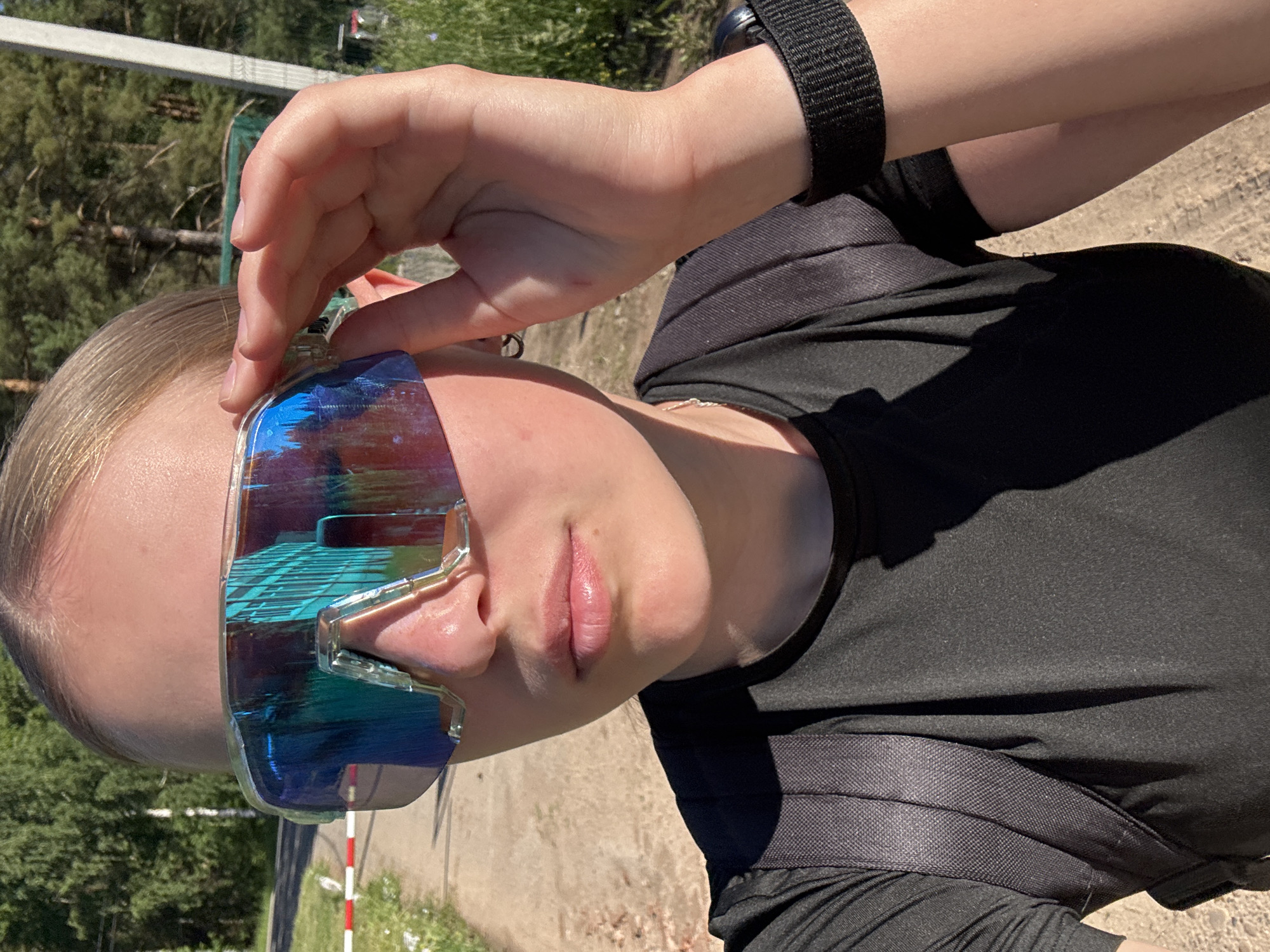 A young woman is taking a selfie outdoors, wearing large, iridescent sport sunglasses. She is looking directly at the camera and is holding the edge of the sunglasses with her right hand. She is wearing a black t-shirt with a backpack on her shoulders. The background shows a dirt path, trees, and a white and red striped pole. The lighting suggests it is daytime with the sun shining. The scene appears to be a rural or park-like setting, possibly on the outskirts of the city of Chaikovsky, Russia, given the natural environment.